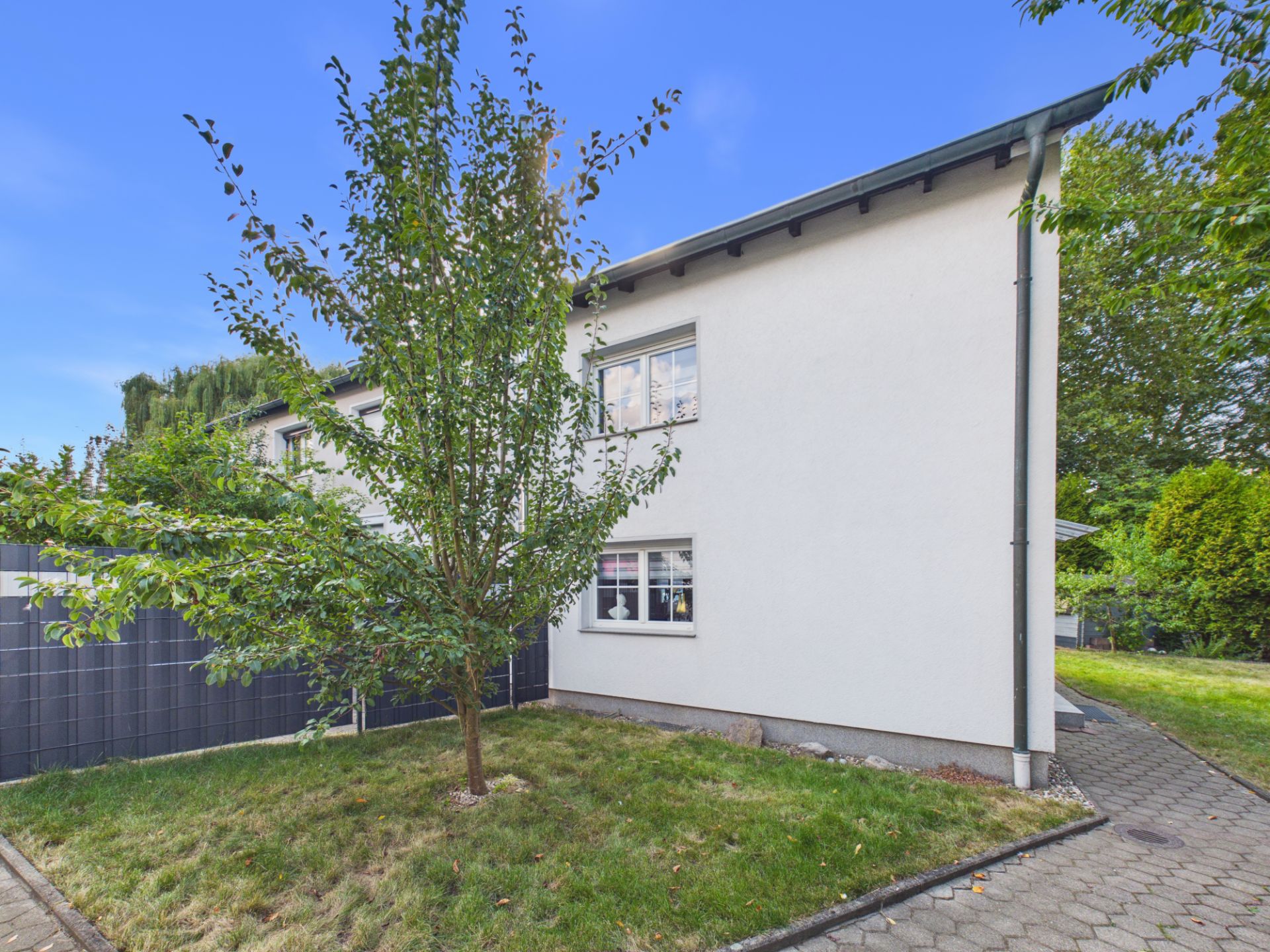 Weiße Hausfassade mit zwei Fenstern und Garten mit kleinem Baum im Vordergrund.