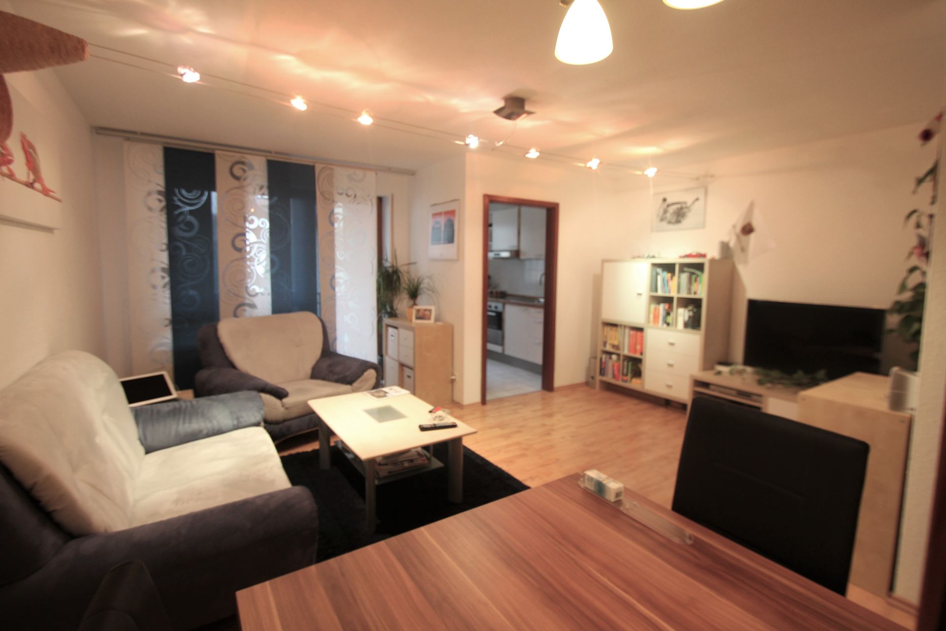 Wohnzimmer mit Sofa, Couchtisch, Fernseher, Regal und Essbereich, Blick zur offenen Küche.