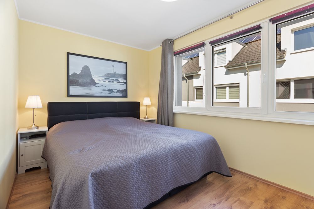 Schlafzimmer mit großem Bett, zwei Nachttischen und Fenstern mit Blick auf andere Häuser.