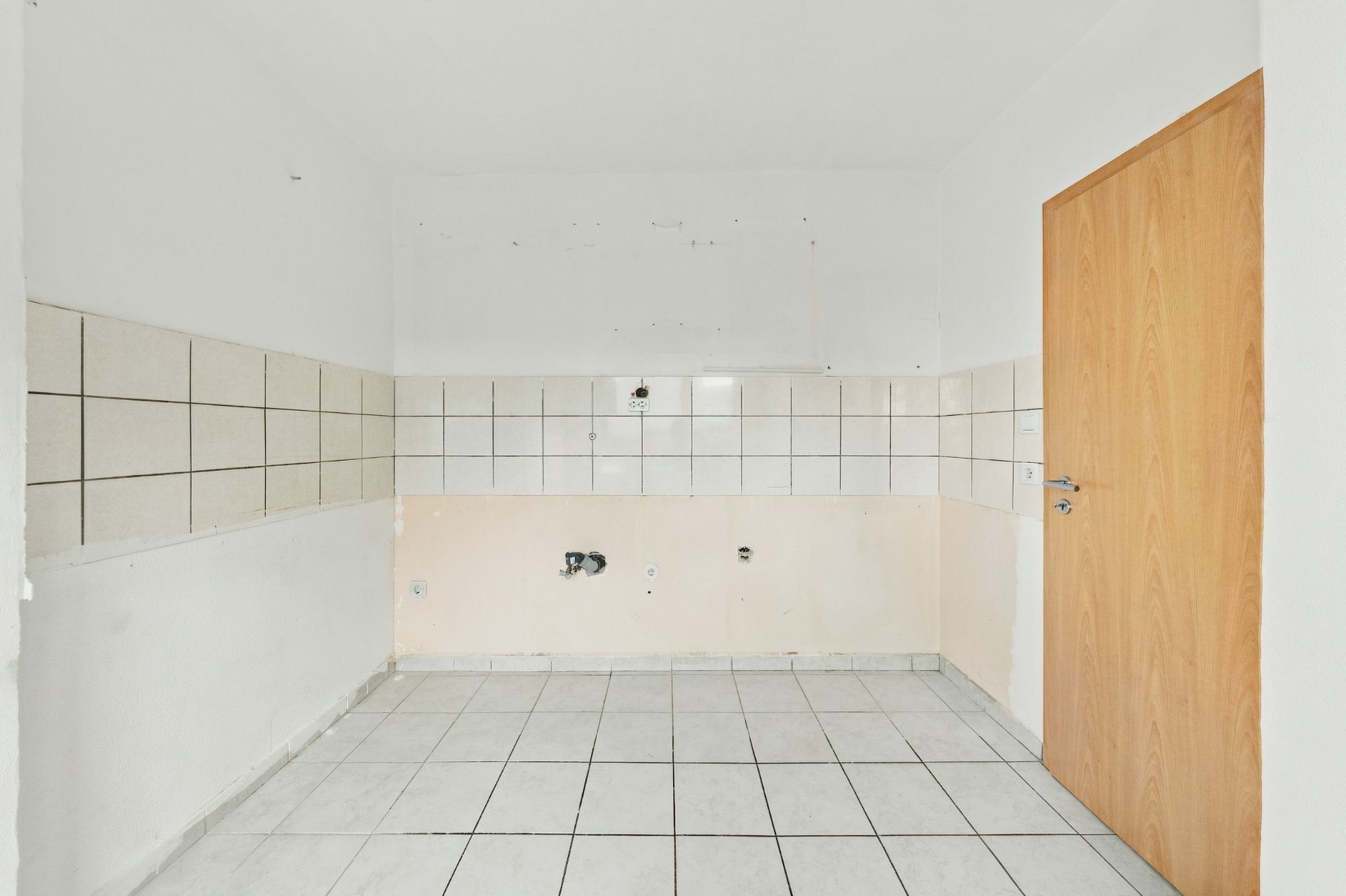 Leerer, gefliester Raum mit weißen Wänden und Holztür, Anschlüsse an der Wand sichtbar.