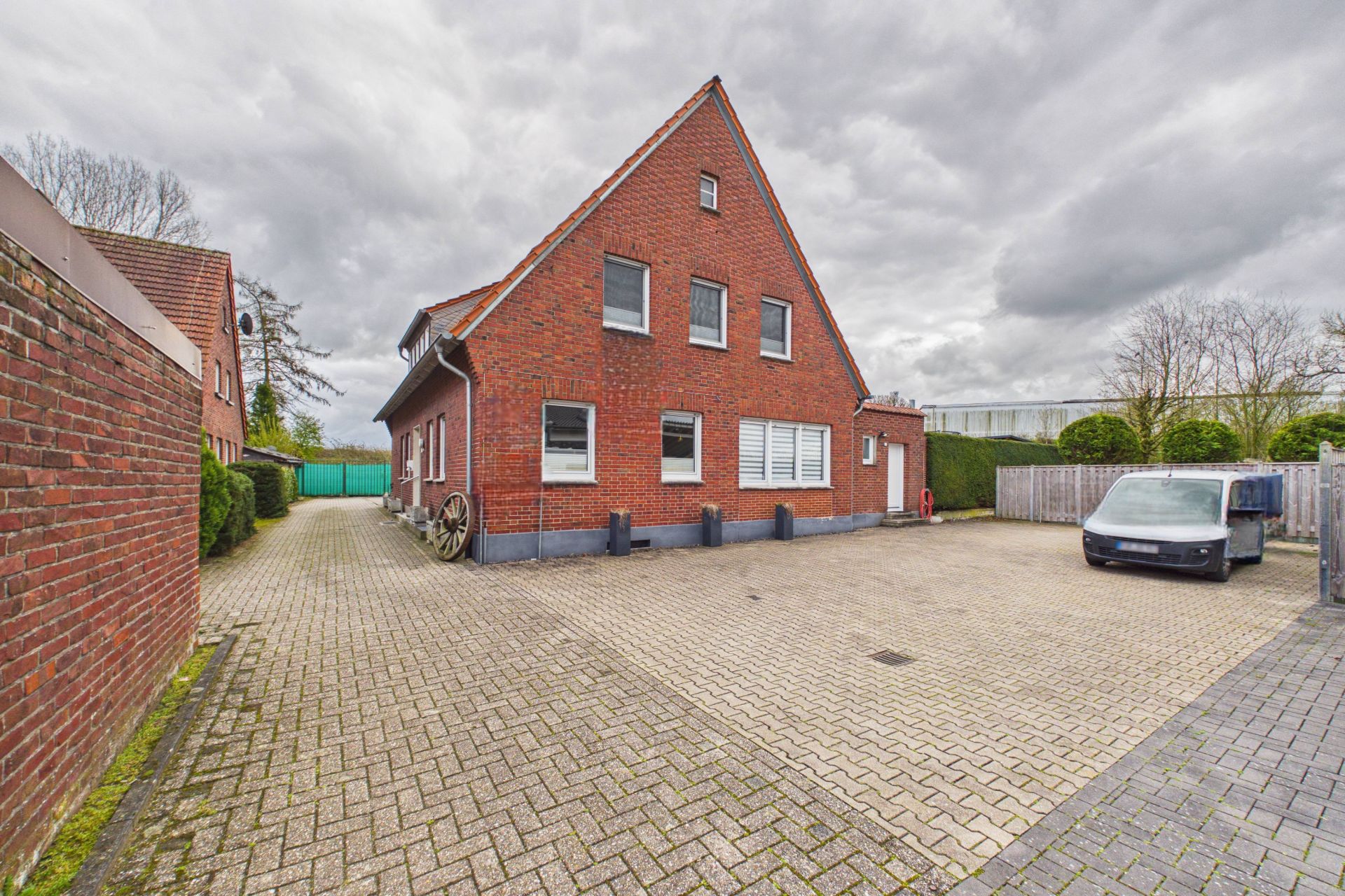 Backsteinwohnhaus mit Satteldach und großem gepflastertem Hof, Auto rechts geparkt.