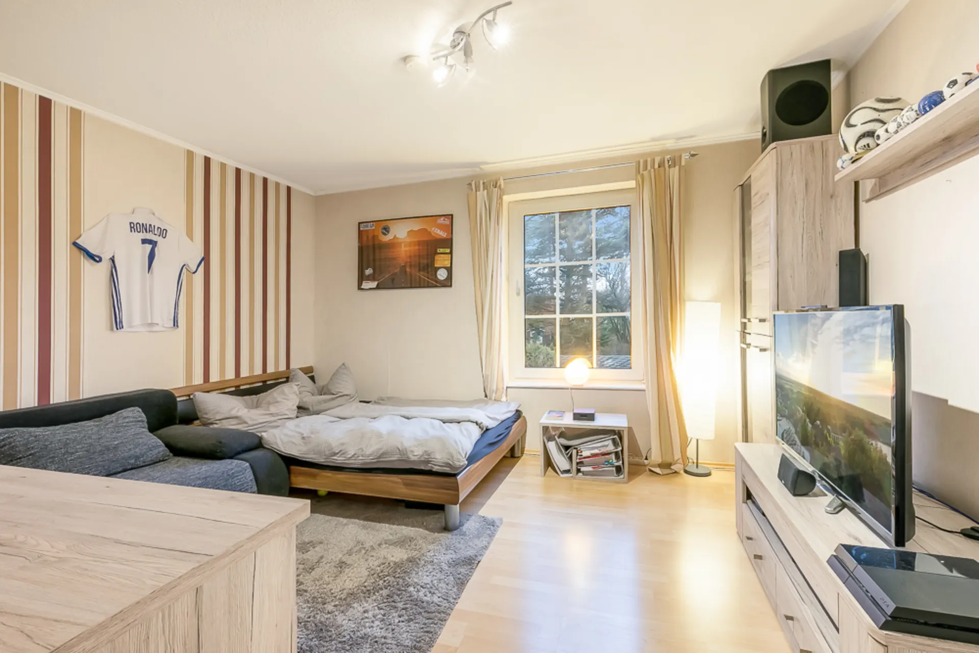 Helles Schlafzimmer mit Bett, Sofa, Fernseher und großem Fenster mit Vorhängen.