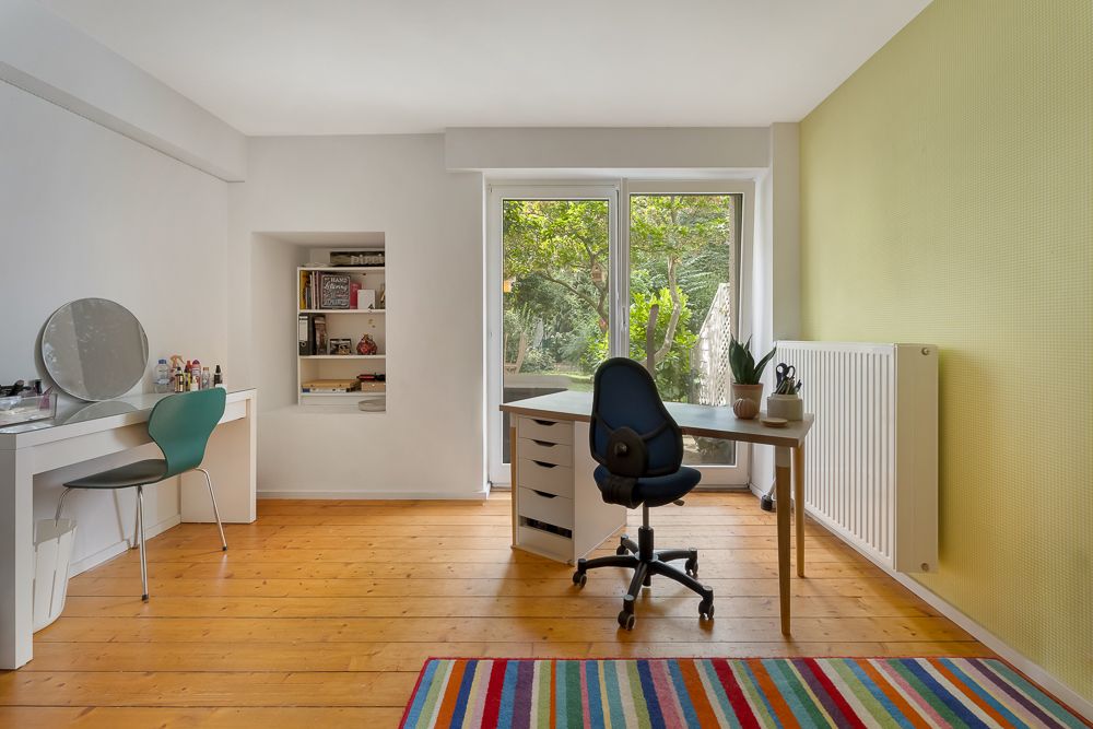 Helles Arbeitszimmer mit Schreibtisch, Stuhl, Regal, Schminktisch und buntem Teppich am Fenster.