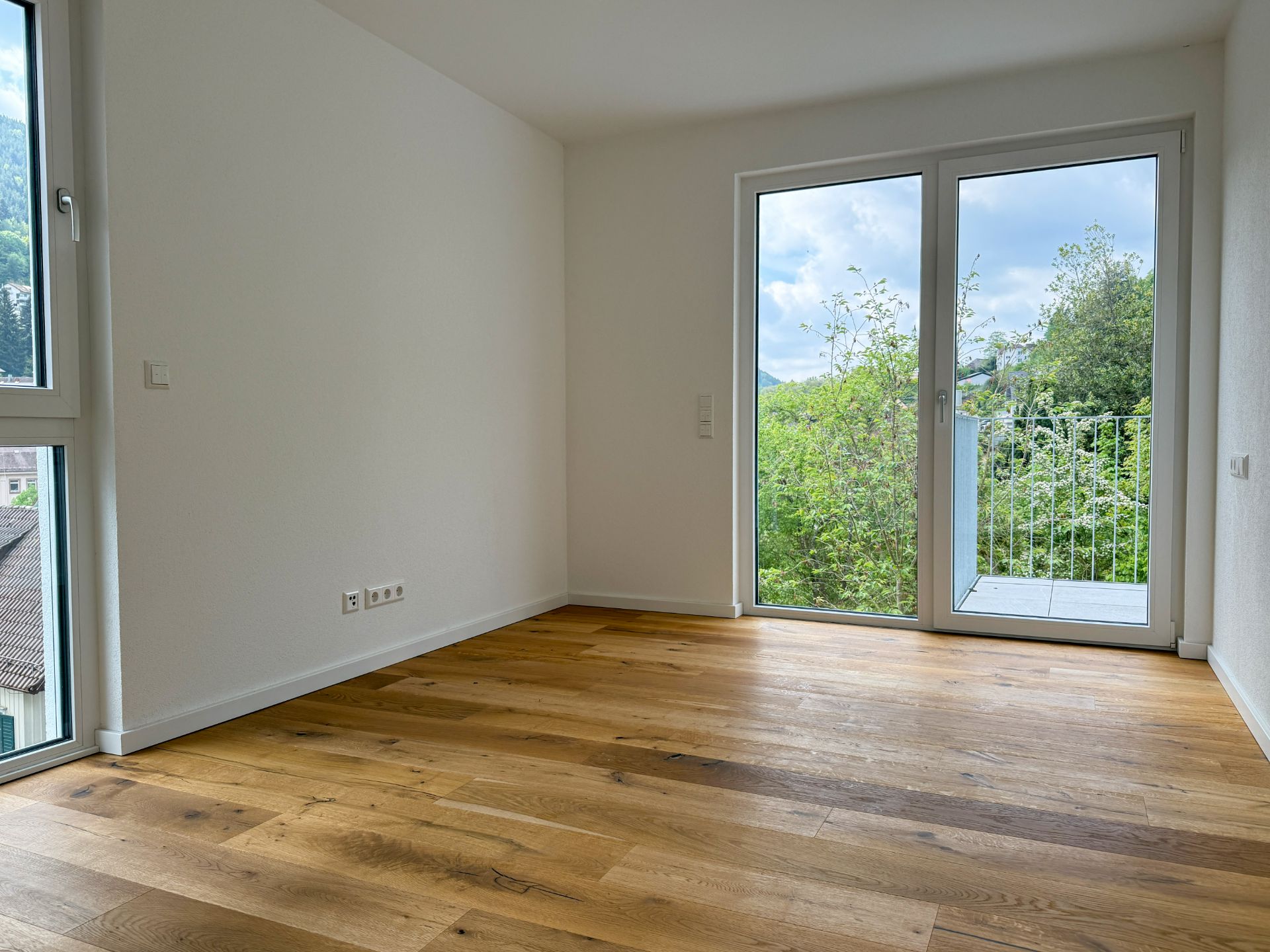 Leeres Zimmer mit Holzboden, weißen Wänden und großer Fensterfront mit Tür zum Balkon.