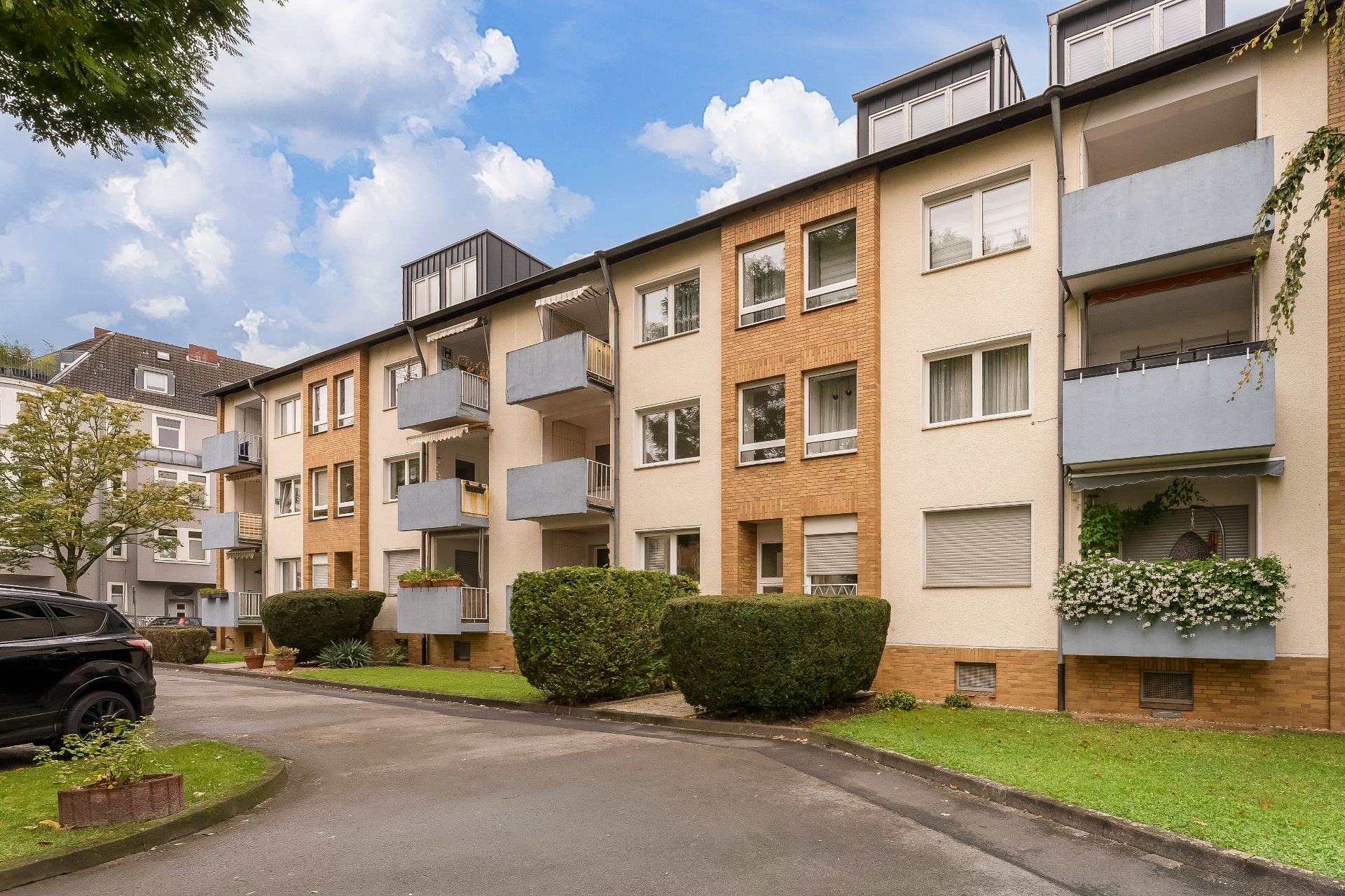 Mehrfamilienhaus mit Balkonen, gepflegtem Vorgarten und parkenden Autos am Weg.