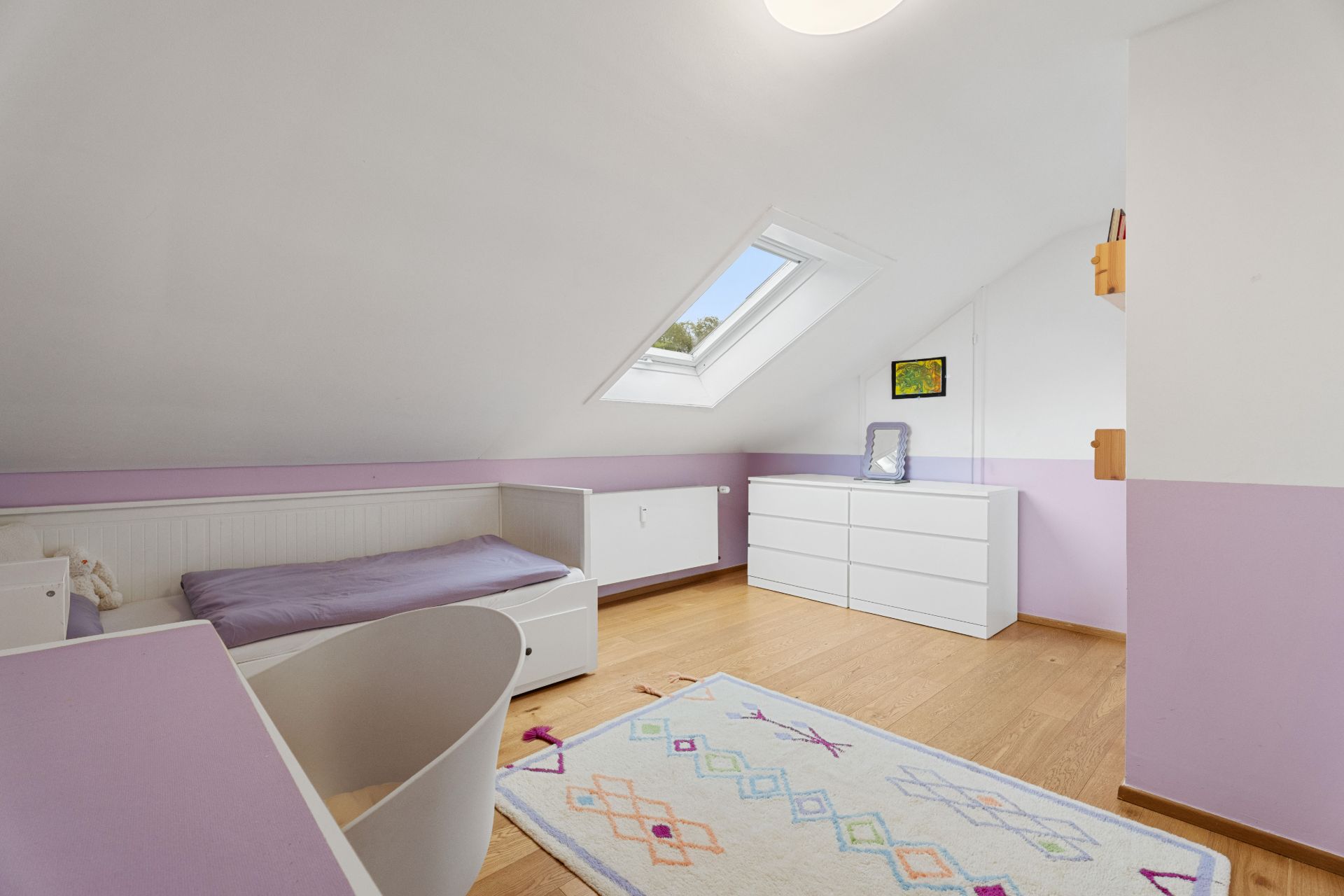 Helles Kinderzimmer mit Schrägdach, Einzelbett, Schreibtisch, Kommode und Teppich.