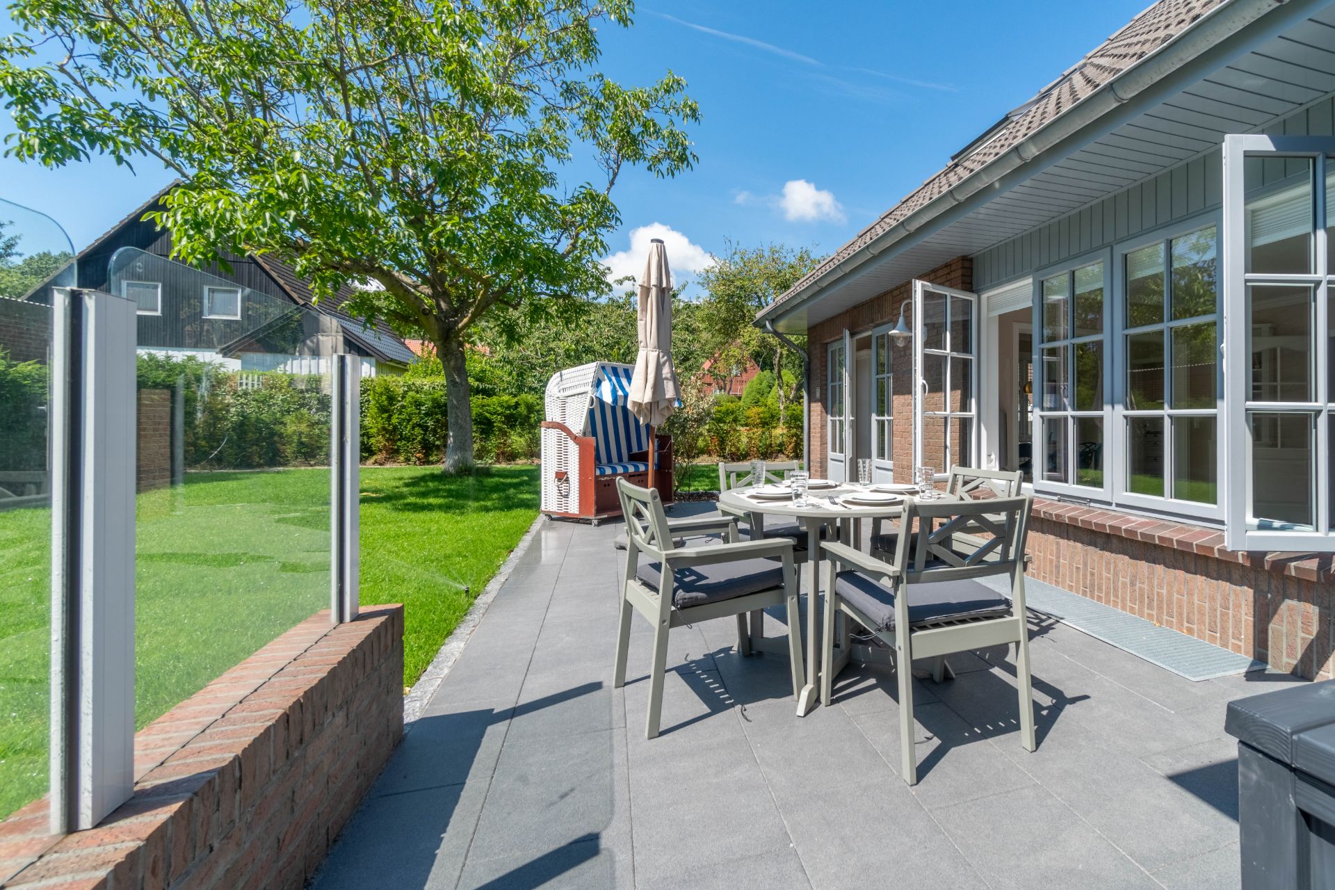 Terrasse mit Esstisch und Stühlen neben Garten, große Fenster und Sonnenschirm sichtbar.
