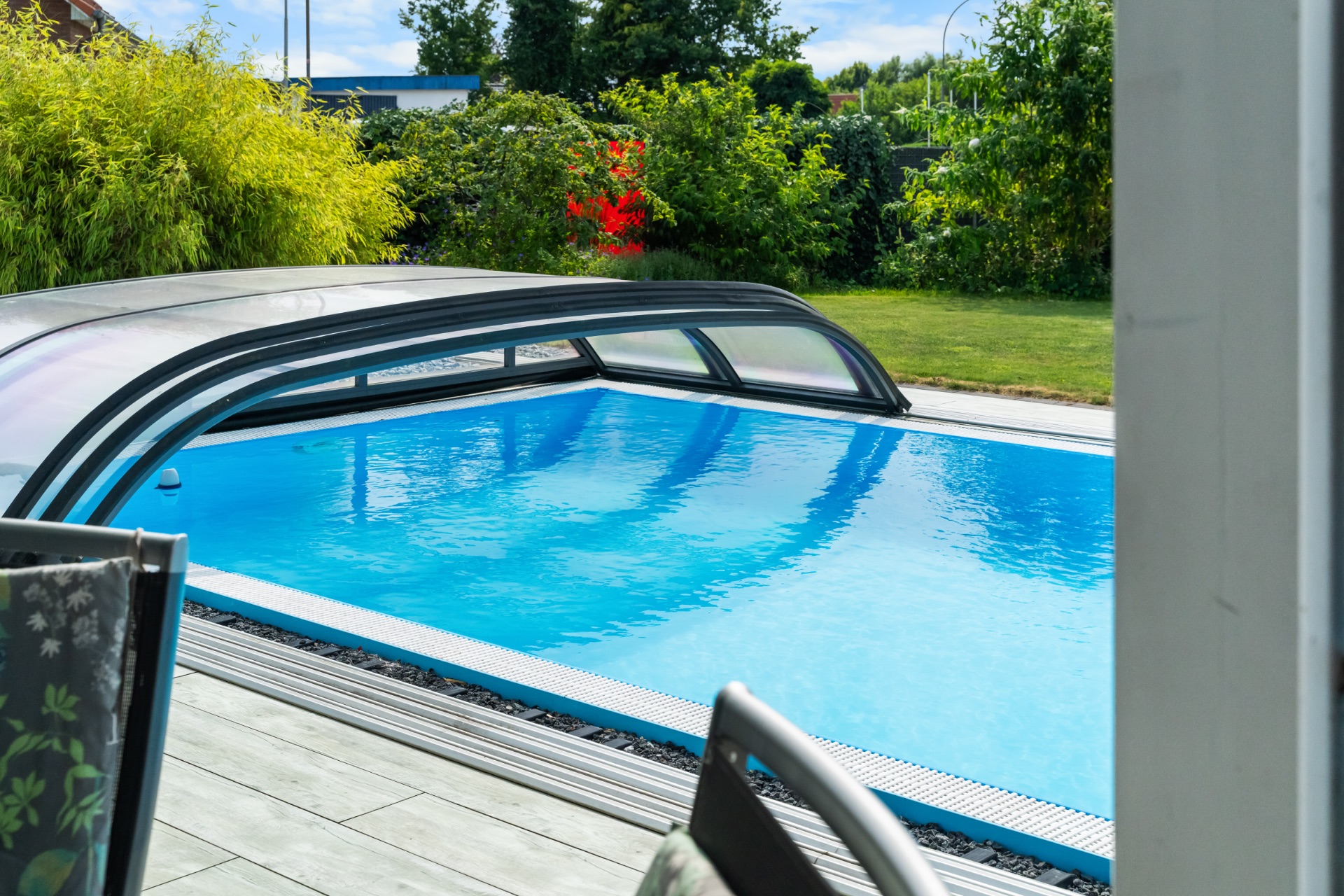 Überdachter Pool im Garten mit umliegendem Grün und Terrasse im Vordergrund.