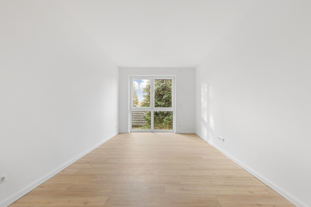 Leerer Raum mit weiß gestrichenen Wänden, Holzfußboden und großem Fenster mit Blick ins Grüne.