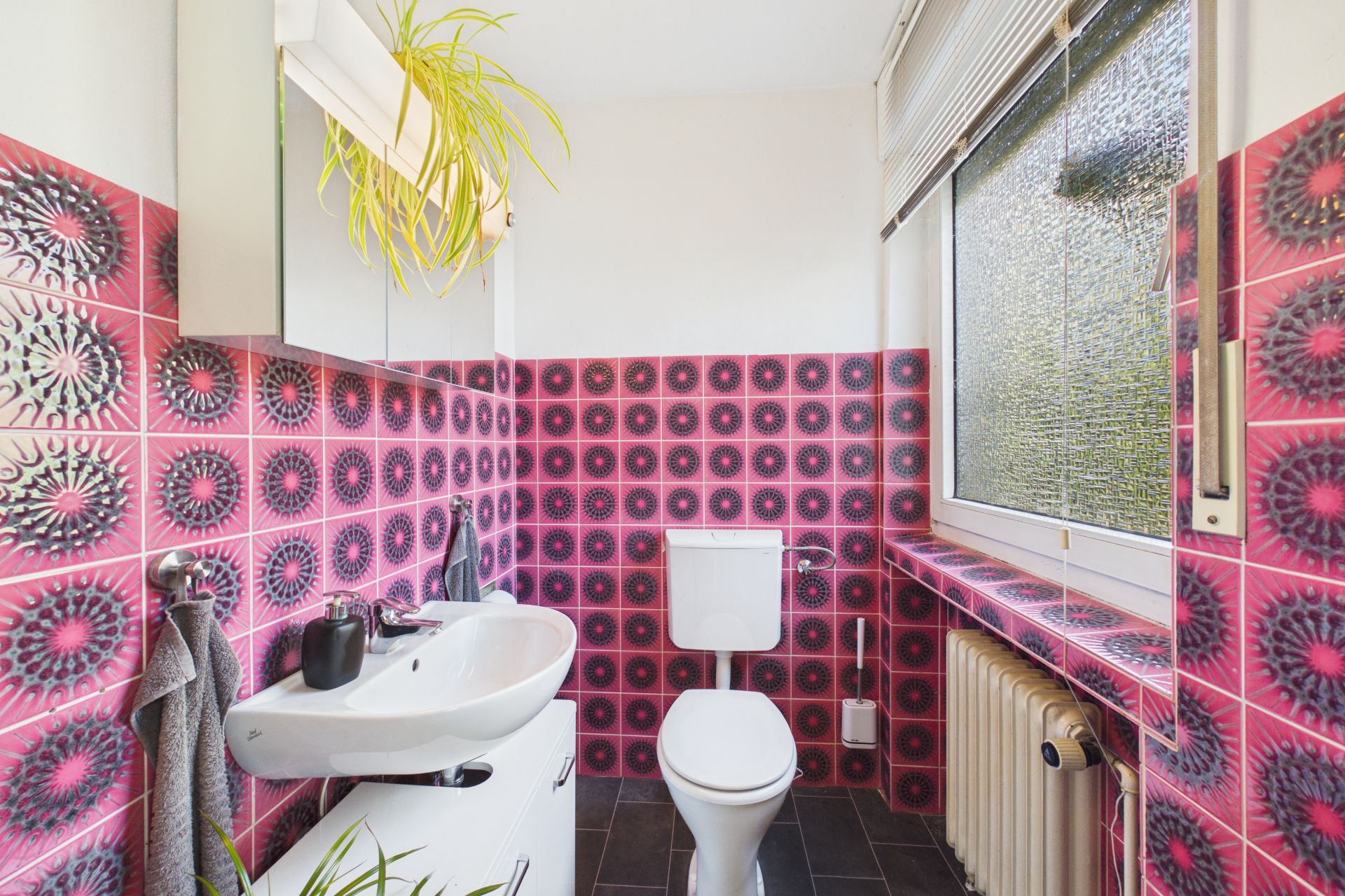 Kleines Badezimmer mit auffälligen pink-schwarzen Fliesen, Waschbecken, Toilette und großem Fenster.
