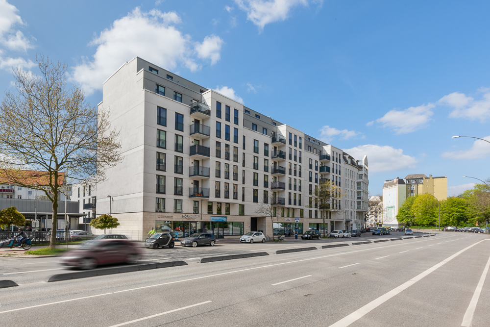 Modernes Wohngebäude mit Balkonen an einer breiten Straße, mehrere Autos parken am Straßenrand.