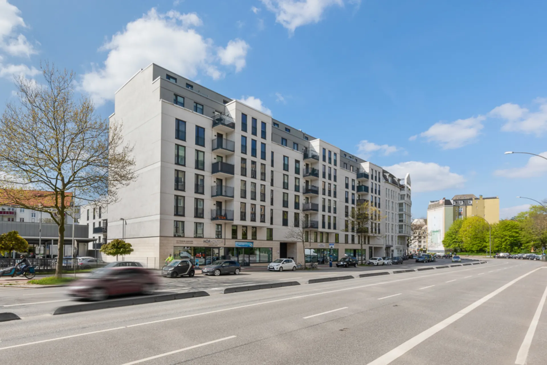 Modernes Wohngebäude mit Balkonen an einer breiten Straße, mehrere Autos parken am Straßenrand.