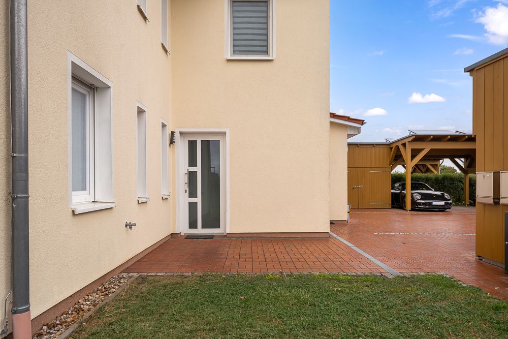 Haus mit hellem Putz, Eingangstür, Rasenfläche und gepflastertem Weg, Carport mit Auto im Hintergrund.