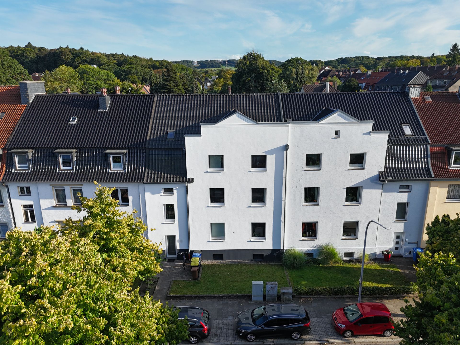 Mehrfamilienhaus mit weißer Fassade und schwarzem Dach, davor geparkte Autos und Bäume.