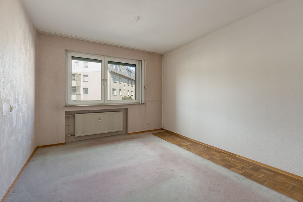 Leeres Zimmer mit zwei Fenstern, Heizkörper und teilweise Teppichboden, teilweise Parkett.
