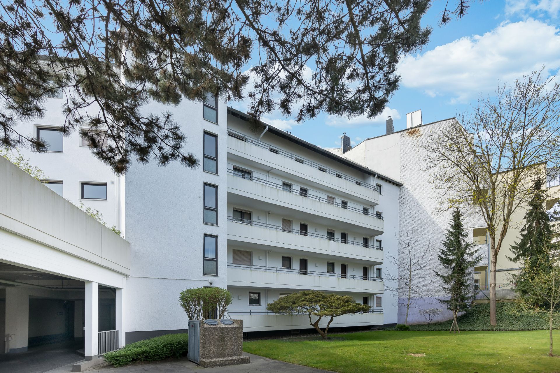 Mehrstöckiges Wohngebäude mit Balkonen und grünem Innenhof, Bäume im Vordergrund.