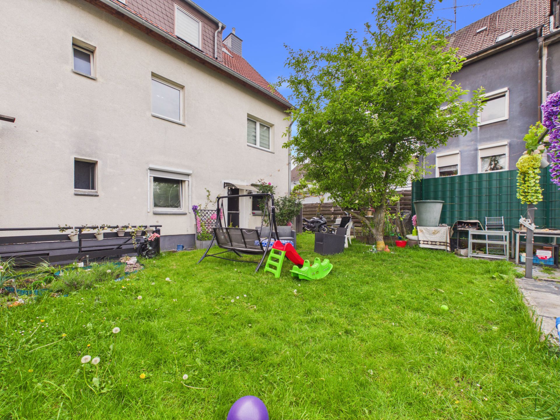 Grüner Garten mit Spielgeräten, Bank, Baum und Rückseite eines Wohnhauses.