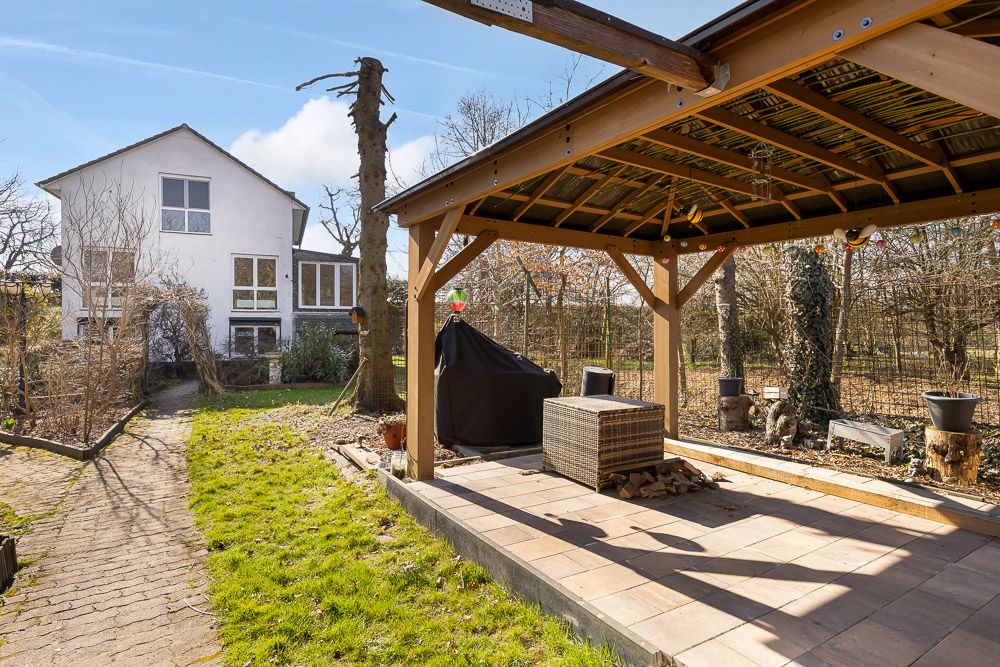 Überdachte Terrasse mit Sitzmöbeln im Garten, Weg führt zum weißen Wohnhaus im Hintergrund.