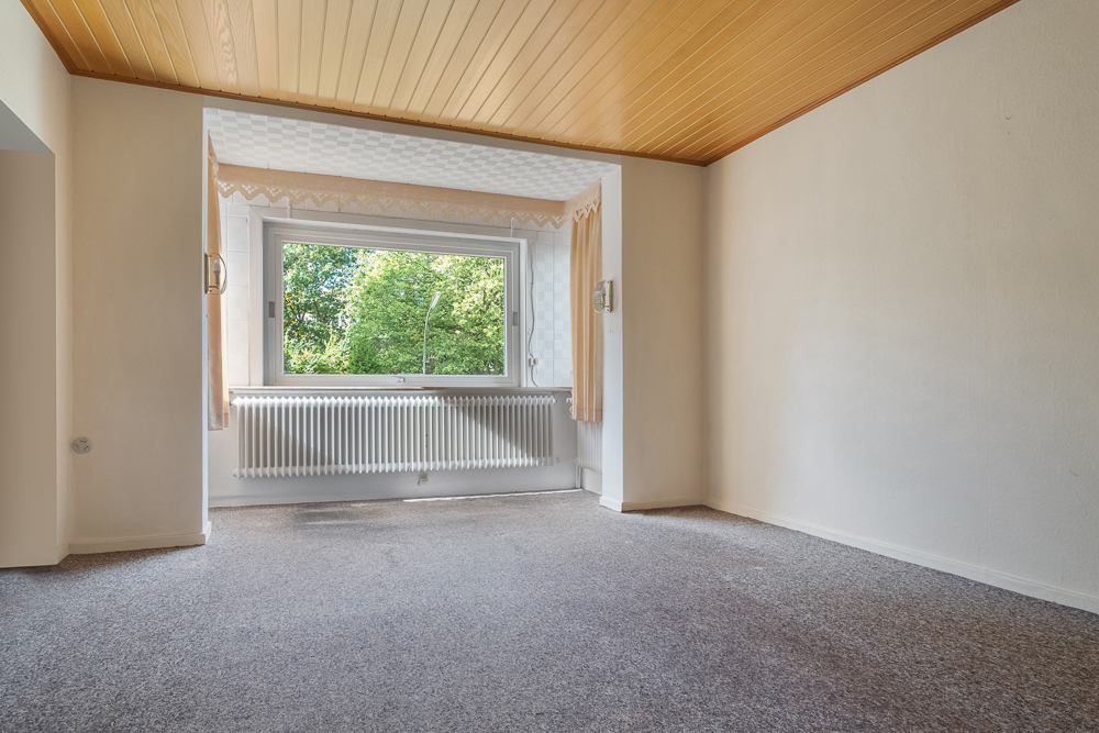 Leeres Zimmer mit Teppichboden, großem Fenster, Heizkörper und Holzdecke.