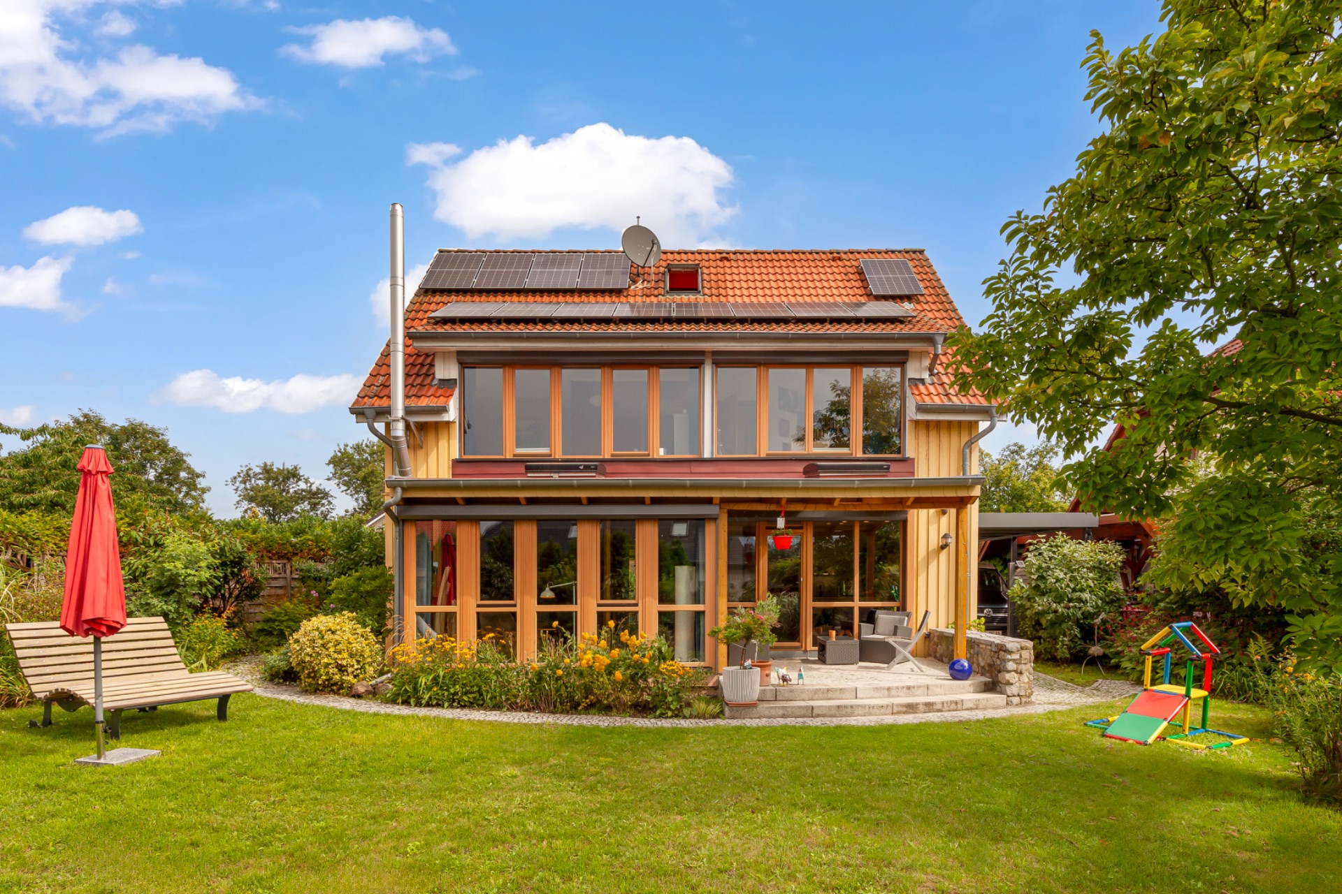 Zweistöckiges Haus mit Wintergarten, Garten, Gartenbank und Kinderspielplatz im Vordergrund.