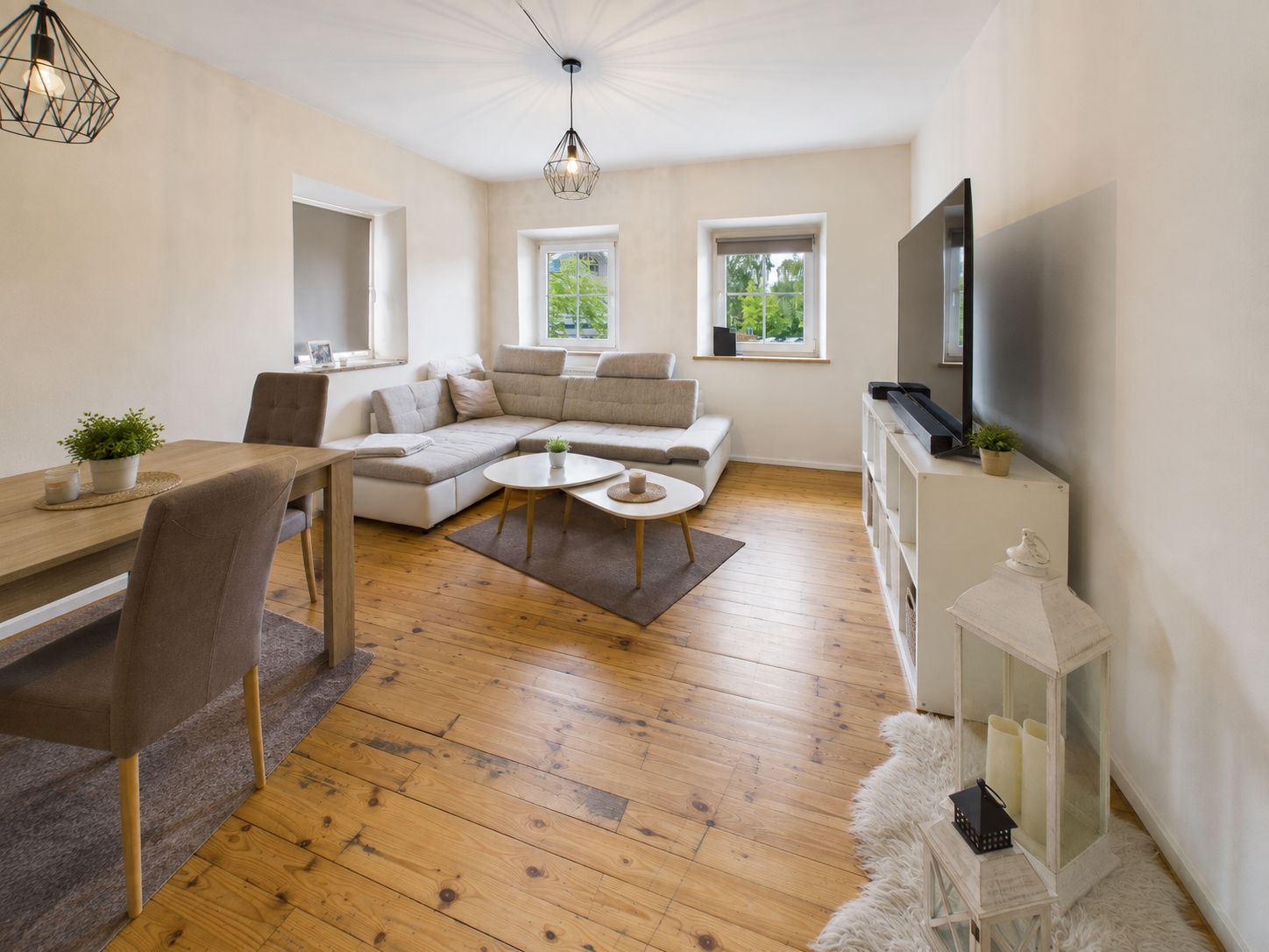 Wohnzimmer mit Holzfußboden, Ecksofa, Esstisch, Fernseher und zwei Fenstern.
