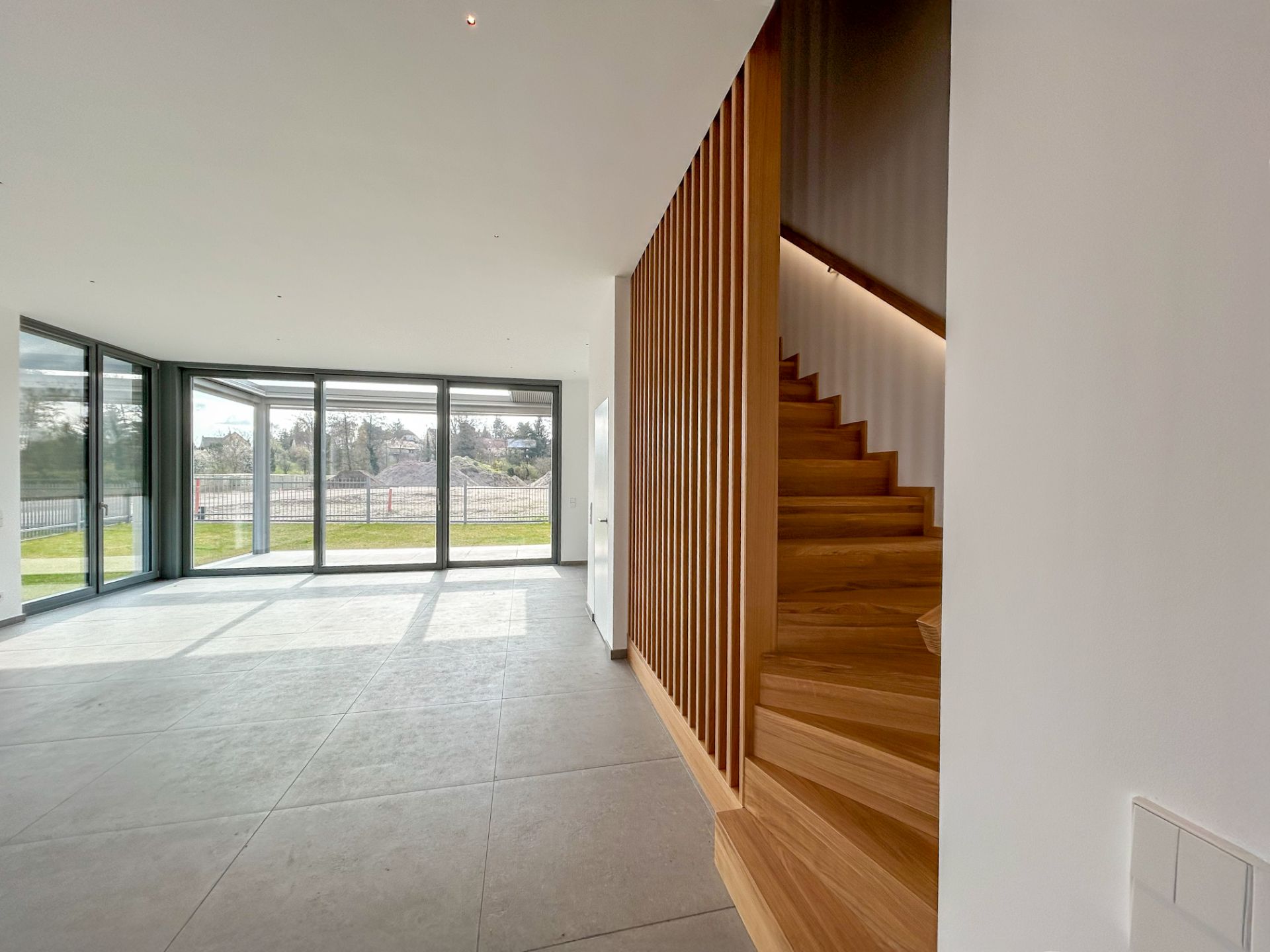 Heller Wohnraum mit großen Fenstern und moderner Holztreppe.