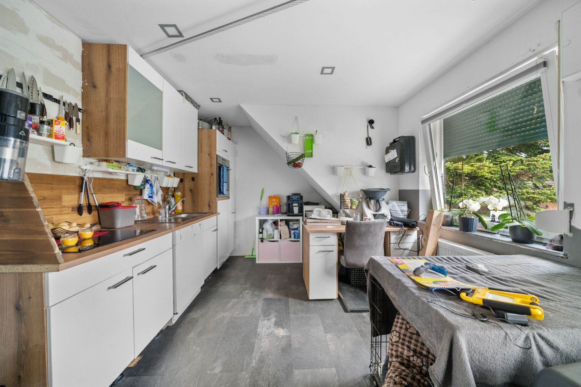 Küche mit Holzmöbeln, Esstisch, großem Fenster und Haushaltsgegenständen auf den Arbeitsflächen.