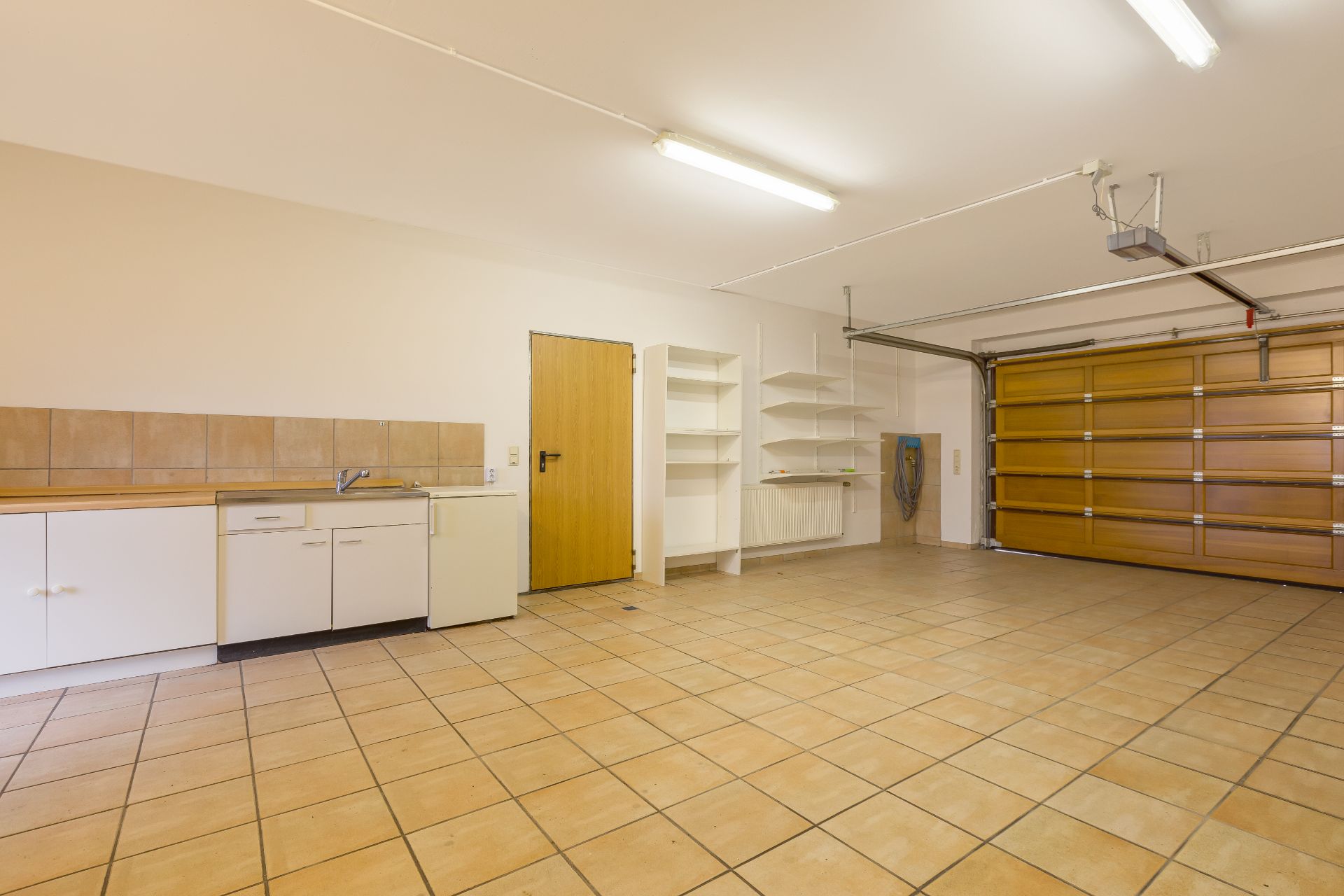 Helle Garage mit Fliesenboden, Holz-Garagentor, Regalen und Waschbecken.