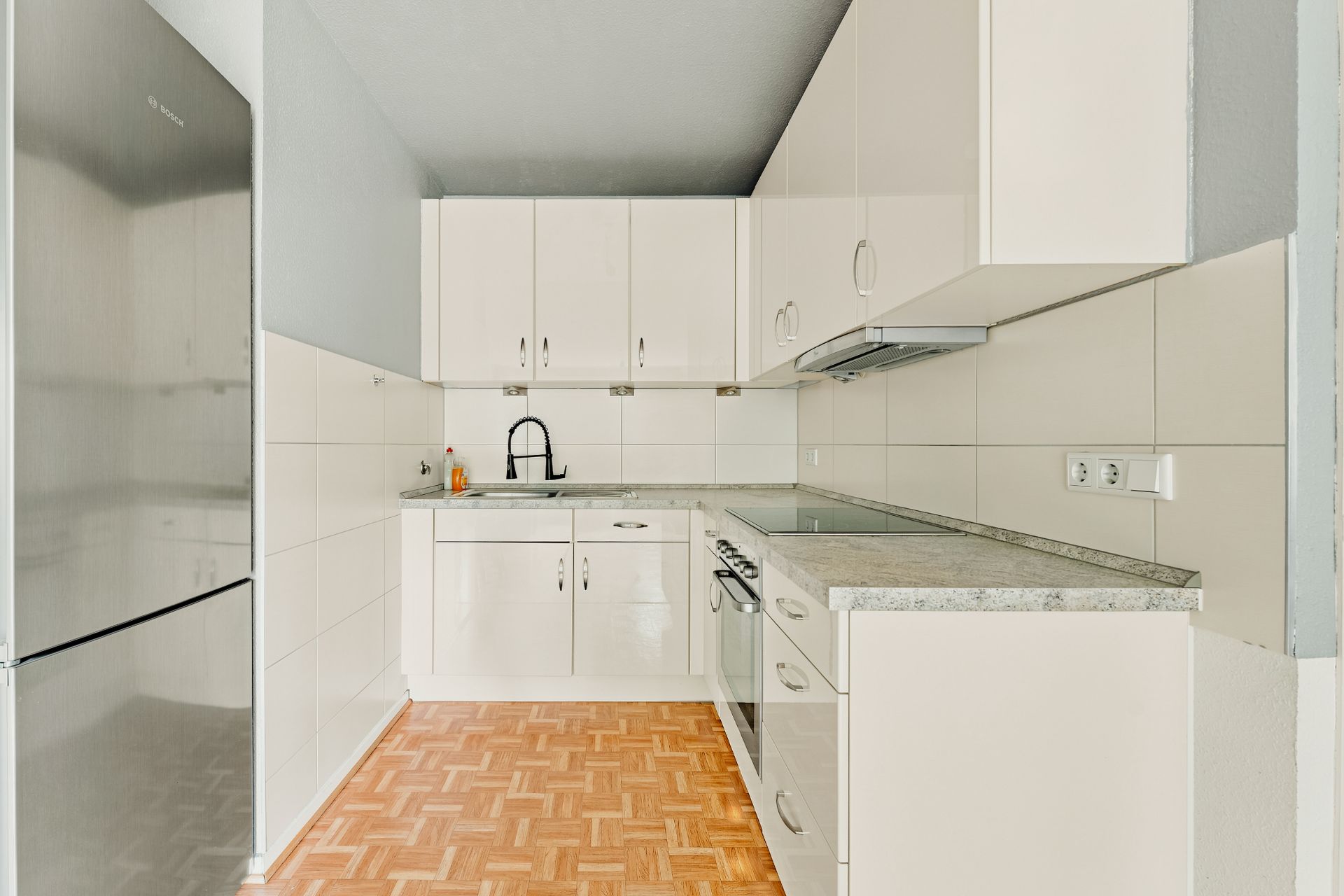 Helle Einbauküche mit weißen Schränken, Herd, Spüle und großem Kühlschrank auf Parkettboden.