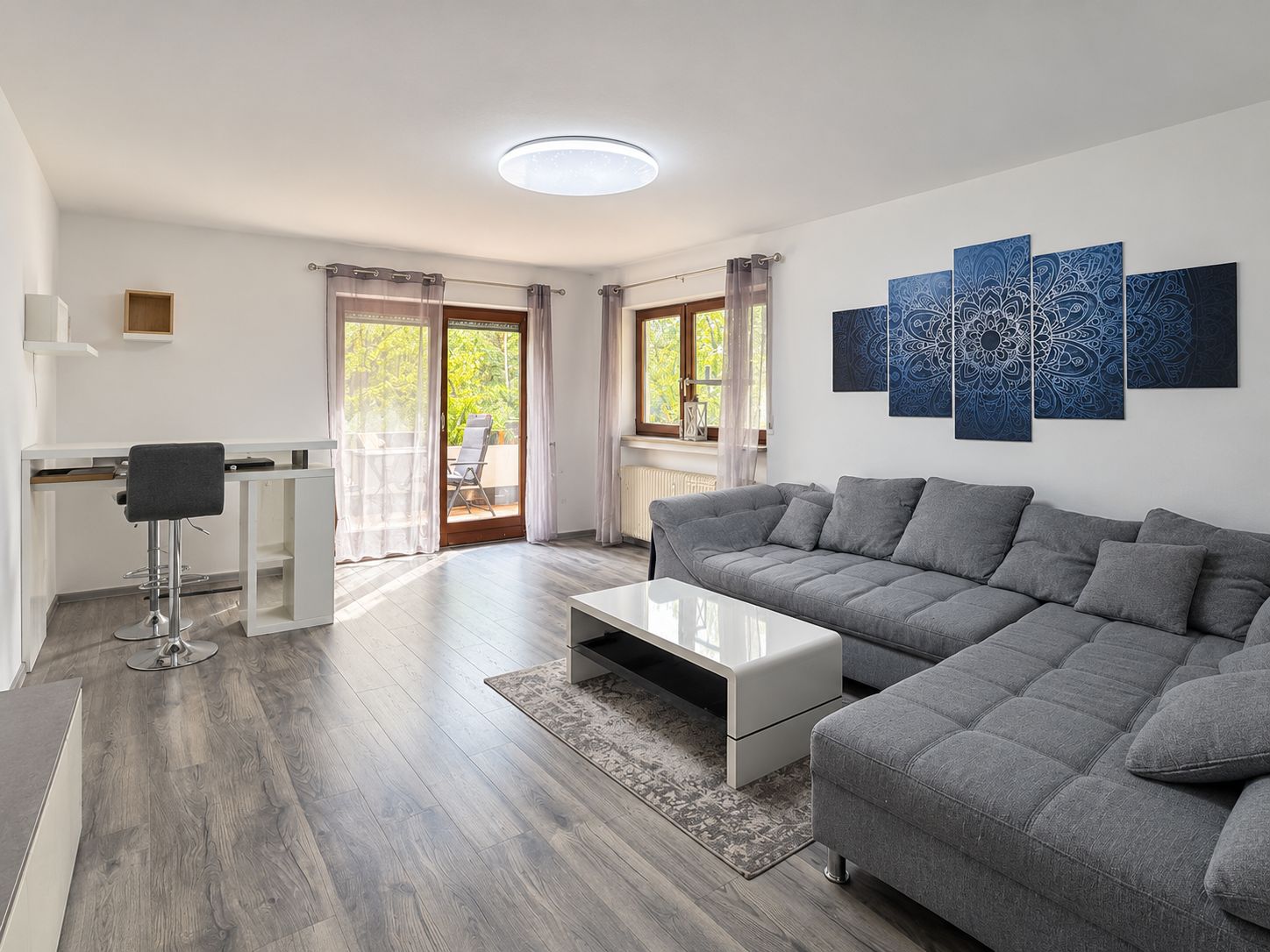 Helles Wohnzimmer mit grauem Ecksofa, Couchtisch, Schreibtisch und Zugang zum Balkon.
