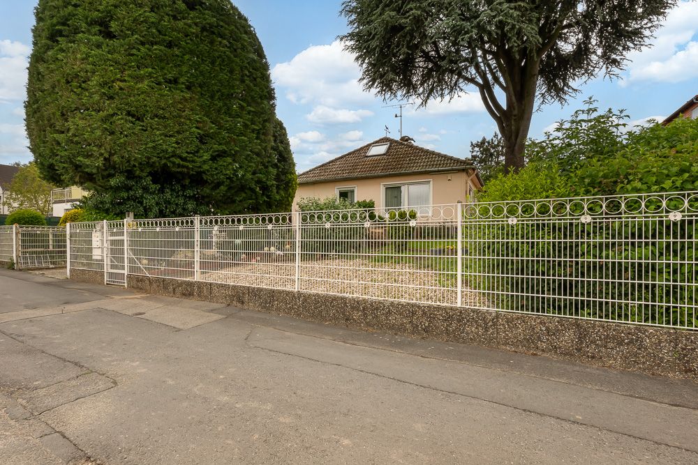 Einfamilienhaus mit Garten, großen Bäumen und weißem Zaun an einer Straße.