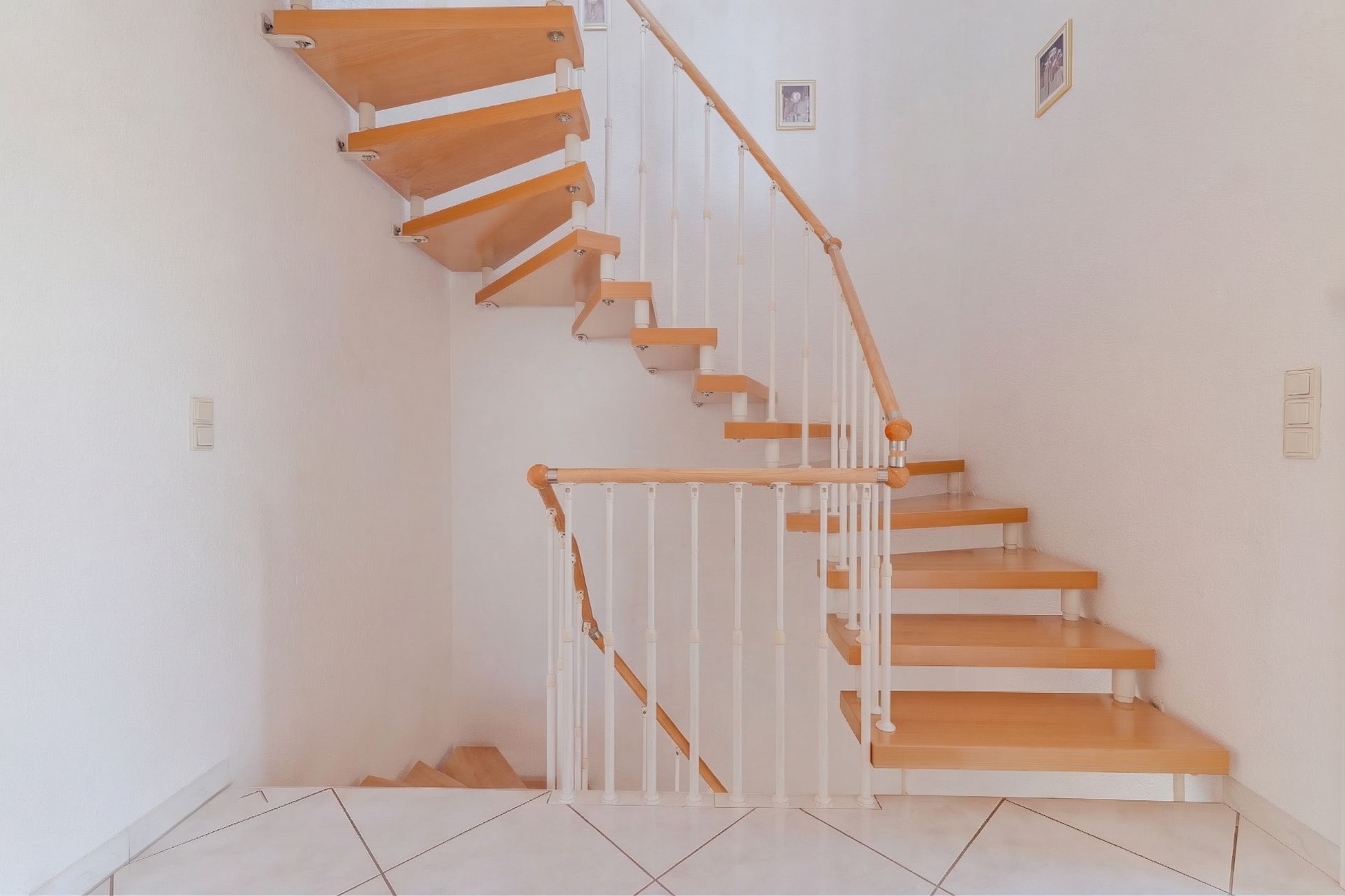 Helle Holztreppe mit weißem Geländer in einem Wohnhaus, weiße Wände und Fliesenboden.