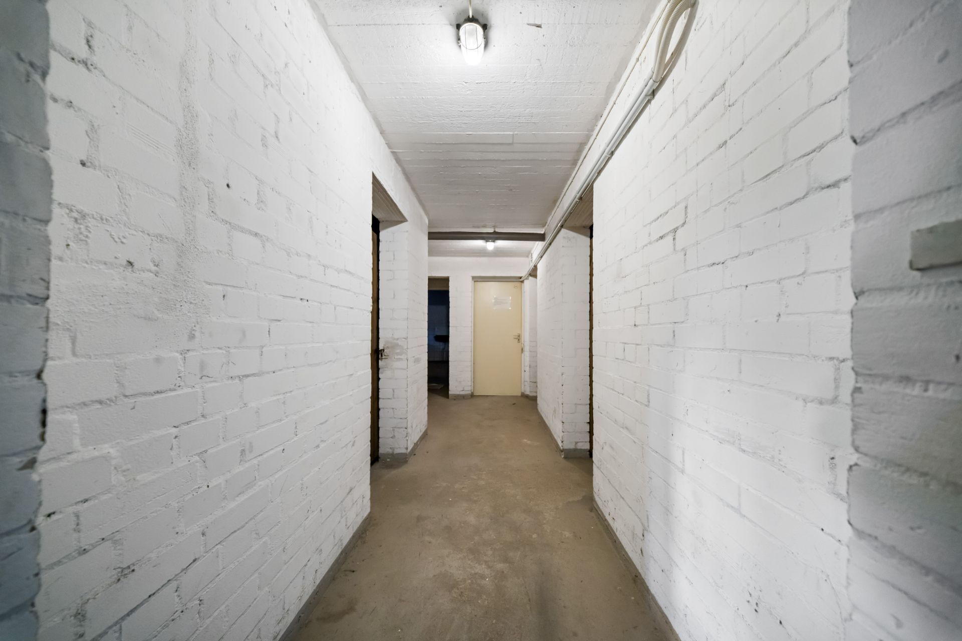 Gang mit weißen Ziegelwänden, vier Türen und Betonboden, Deckenlampe leuchtet.