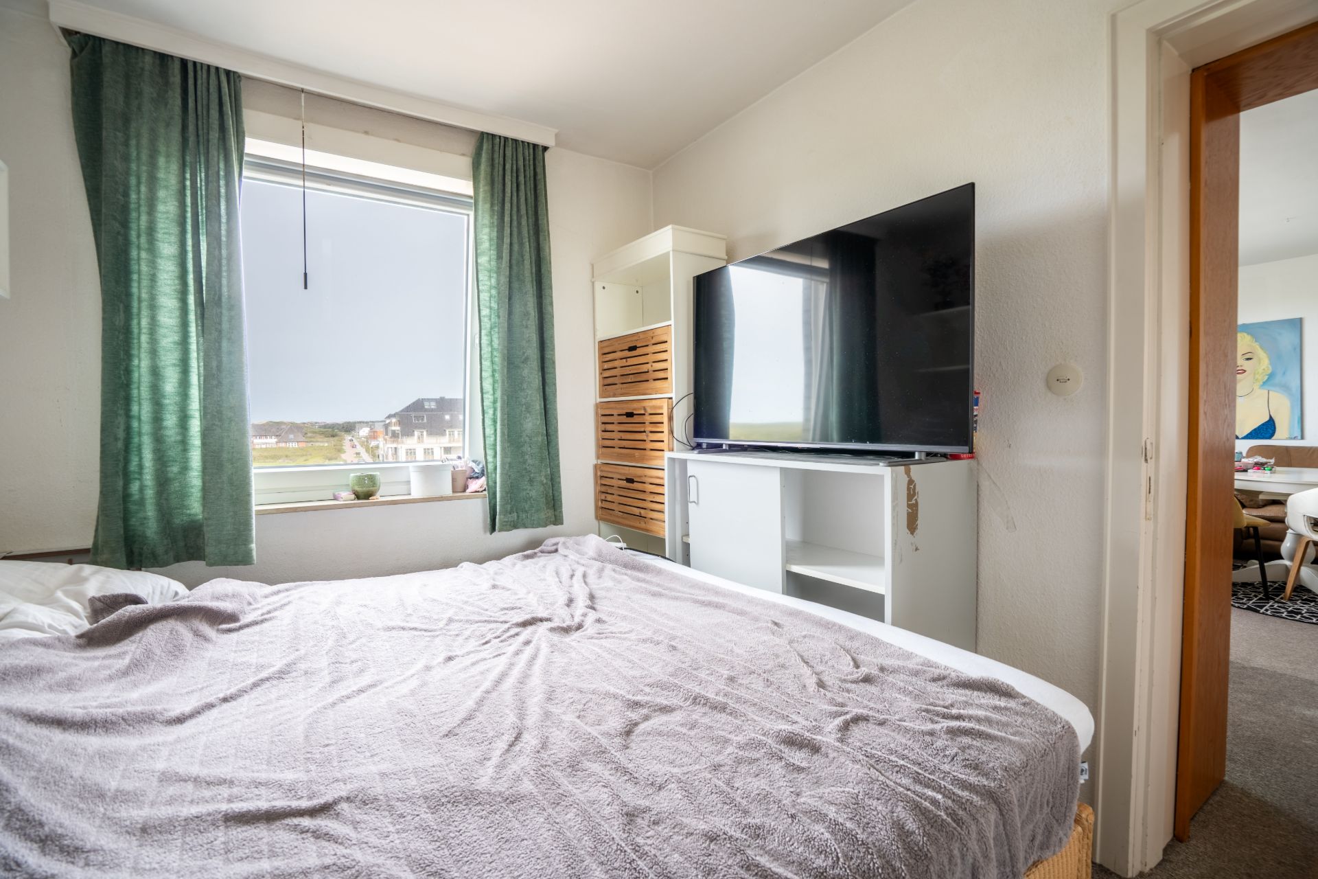 Schlafzimmer mit Bett, Fernseher, Fenster mit grünen Vorhängen und Blick in einen angrenzenden Raum.