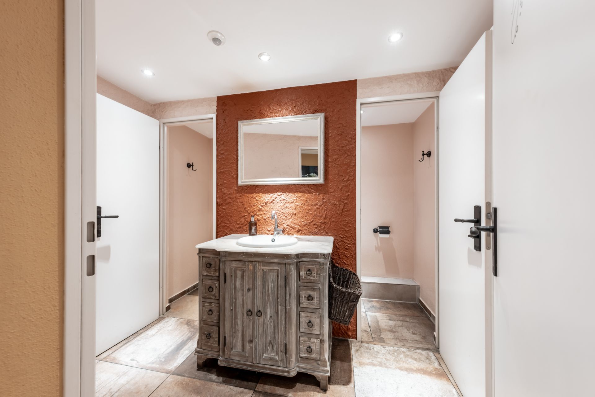 Waschbecken mit Spiegel, Holzmöbel und zwei offene Türen zu separaten Toiletten.