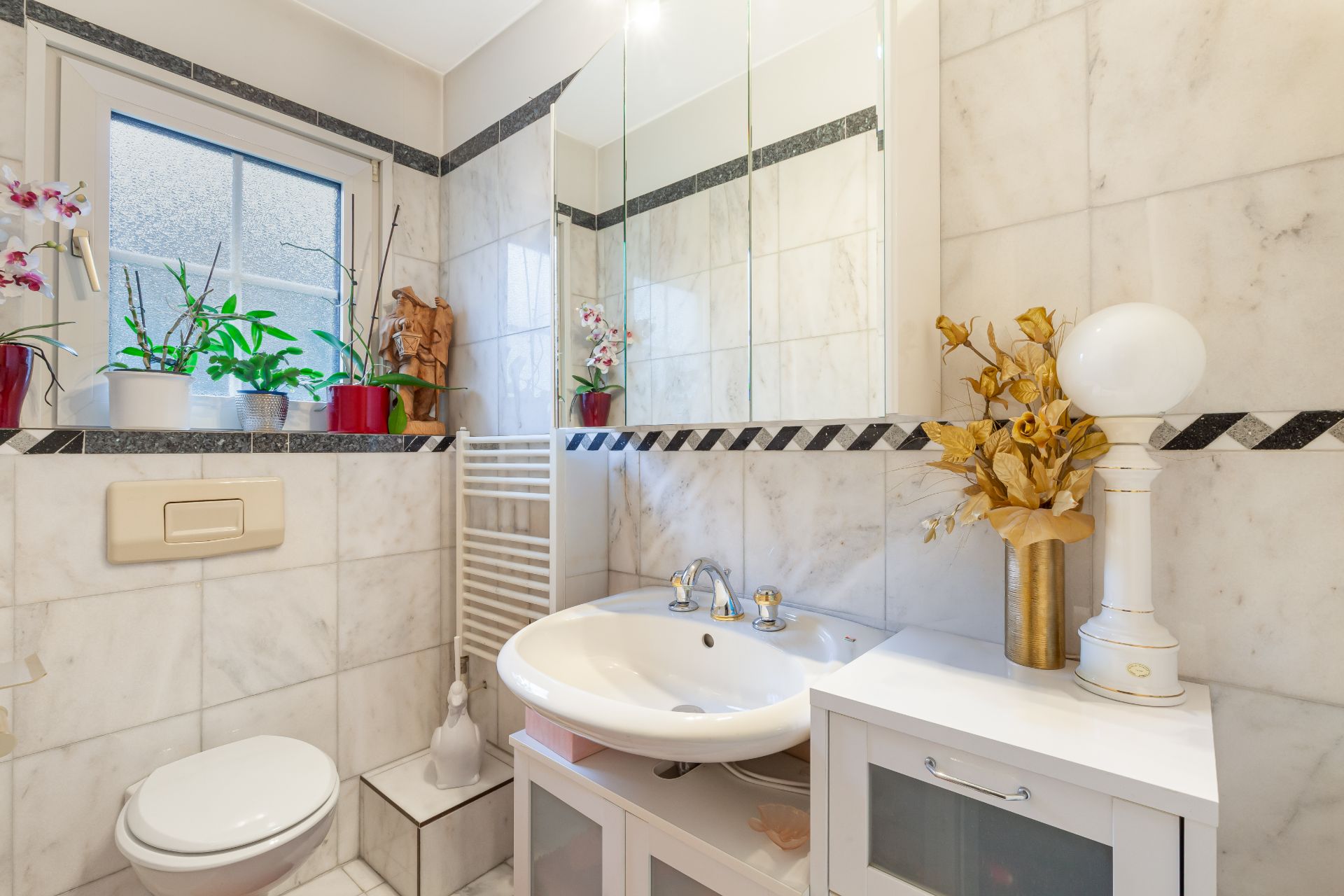 Helles Badezimmer mit Waschbecken, Spiegel, Toilette, Pflanzen und weiß gefliesten Wänden.