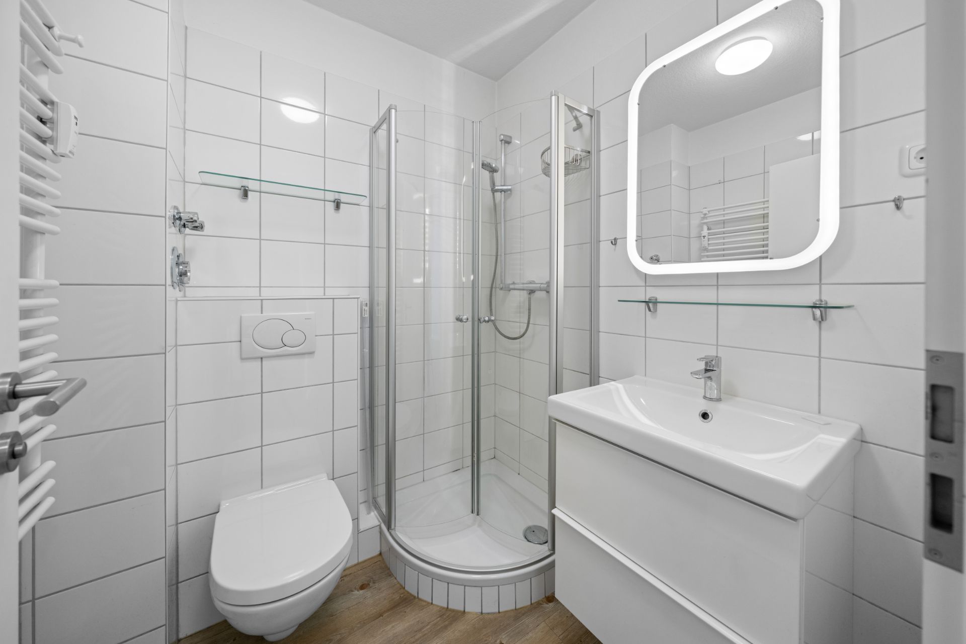 Modernes, weiß gefliestes Bad mit Dusche, WC, Waschbecken und beleuchtetem Spiegel.