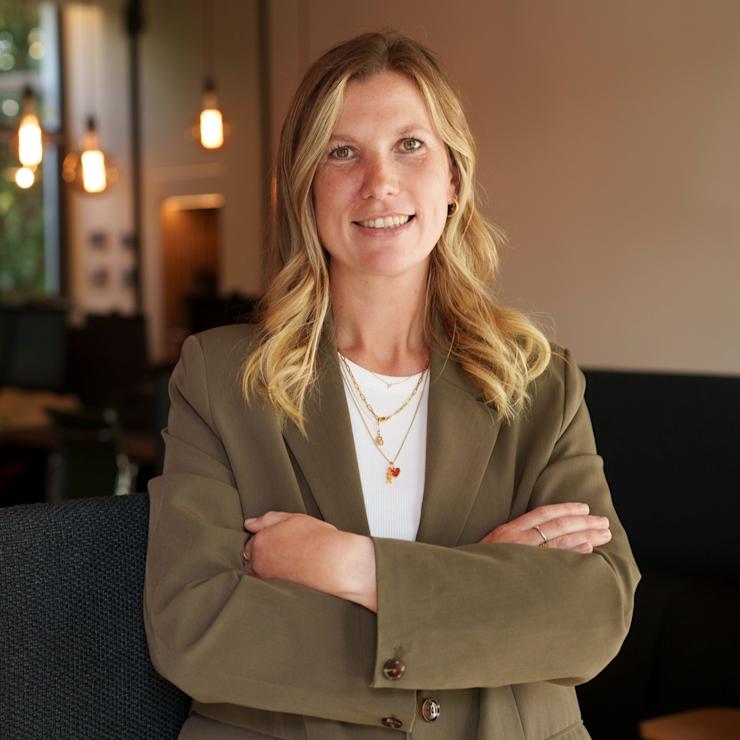 Frau mit blondem Haar und grünem Blazer sitzt mit verschränkten Armen in einem Wohnraum.