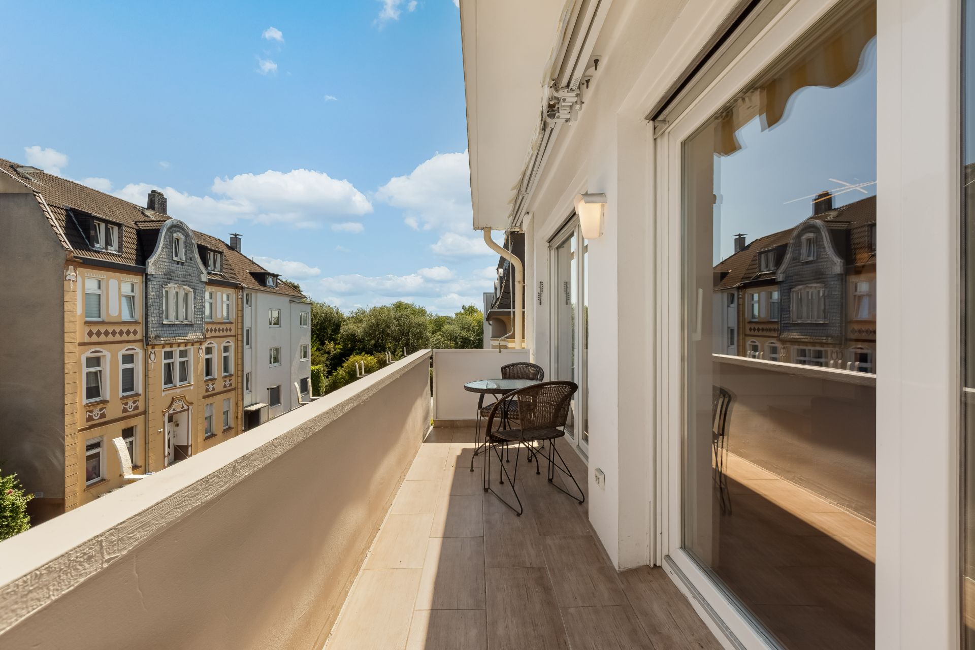 Balkon mit zwei Stühlen und einem Tisch, Blick auf Nachbarhäuser und blauen Himmel.