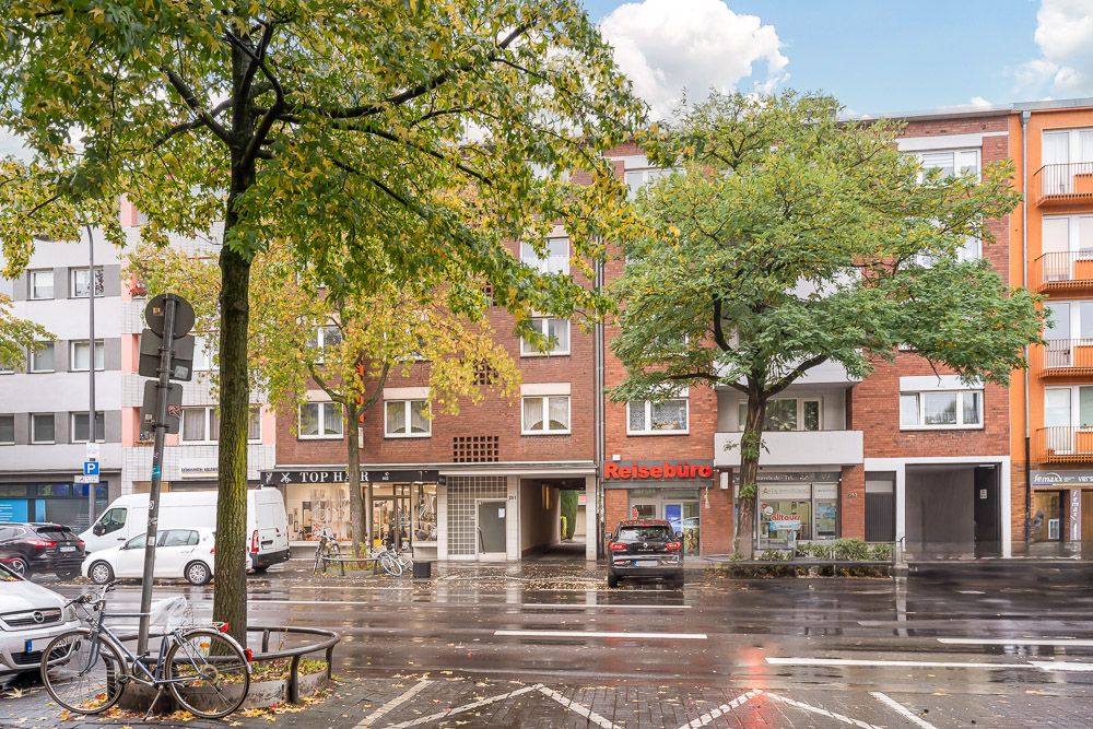 Straßenszene mit Wohnhaus, Bäumen, parkenden Autos und Geschäften im Erdgeschoss.