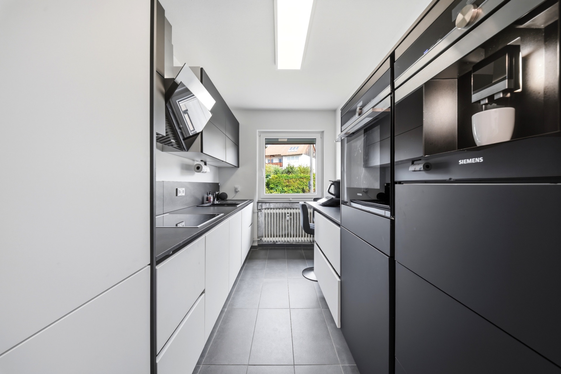 Moderne, schmale Küche mit grauen Schränken, Geräten und Fenster mit Blick ins Grüne.