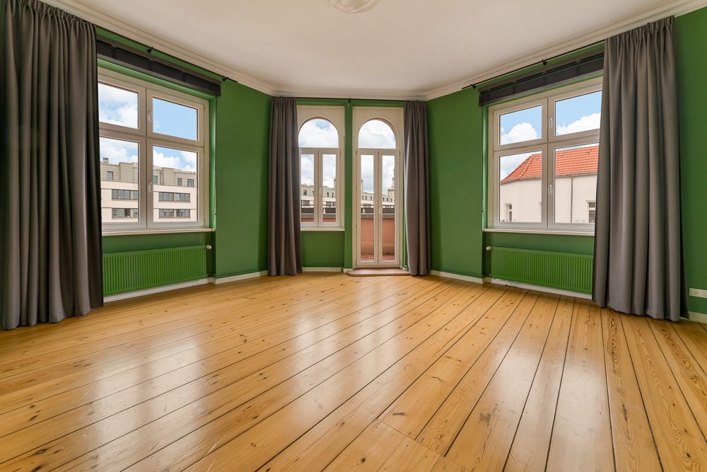 Leerer Raum mit grün gestrichenen Wänden, Holzfußboden und drei großen Fenstern mit Vorhängen.