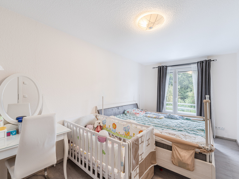 Schlafzimmer mit großem Bett, Babybett und Schminktisch vor einem Fenster mit Vorhängen.