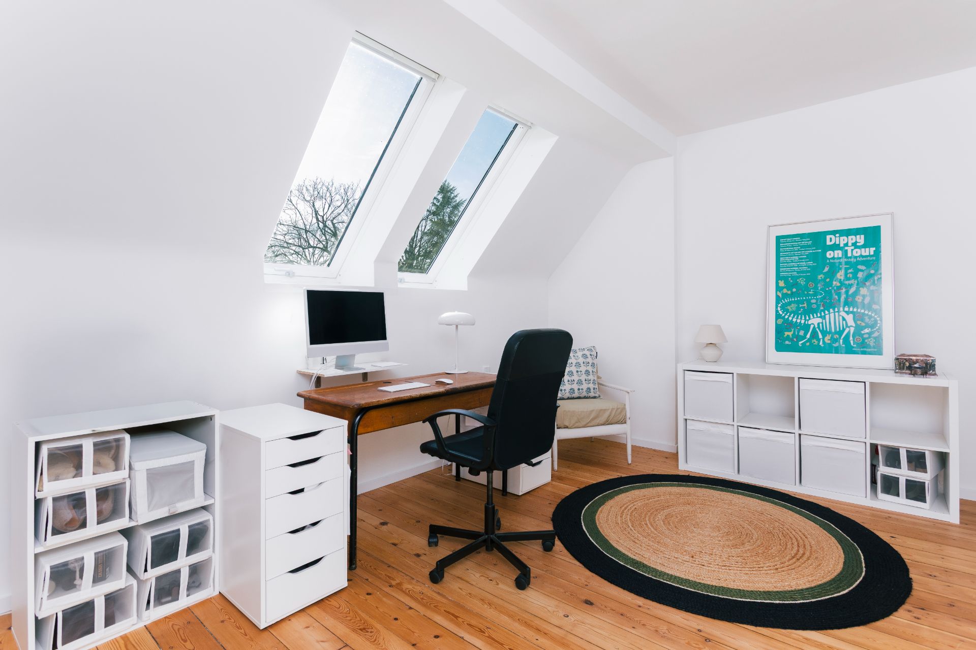 Helles Arbeitszimmer mit Schreibtisch, Stuhl, Regalen und runder Teppich auf Holzfußboden.
