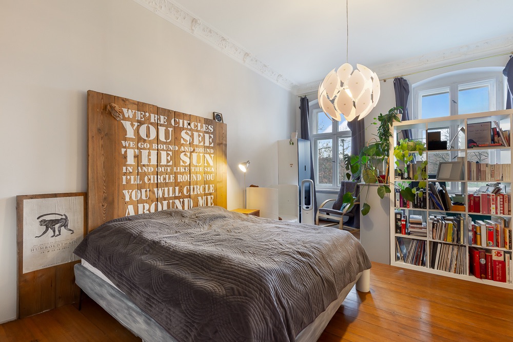 Helles Schlafzimmer mit Doppelbett, großem Bücherregal, Pflanzen und modernem Kronleuchter.
