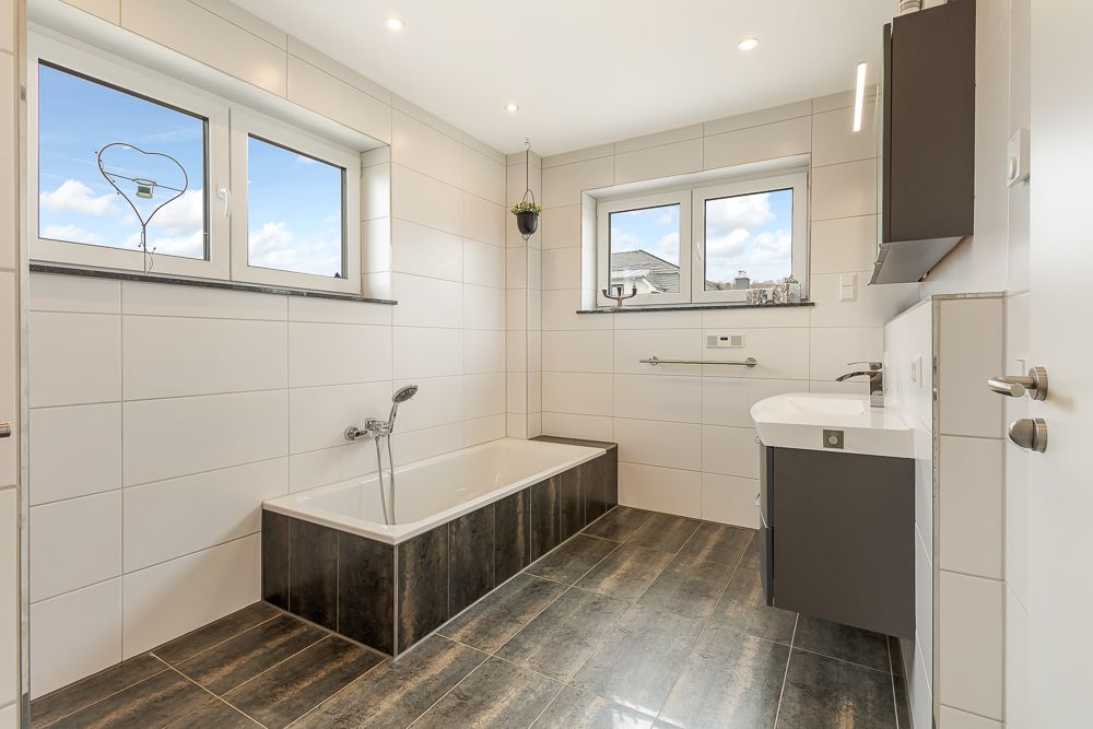 Modernes Badezimmer mit Badewanne, Waschbecken und zwei Fenstern, gefliestem Boden und weißen Wandfliesen.