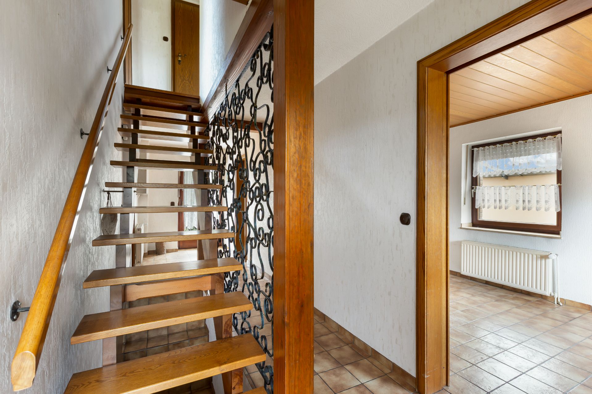 Holztreppe mit Geländer und Flur, angrenzendes Zimmer mit Fenster und Heizkörper sichtbar.