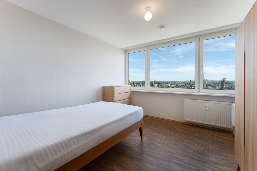 Helles Schlafzimmer mit Einzelbett, Kommode, Kleiderschrank und großem Fenster mit Ausblick.
