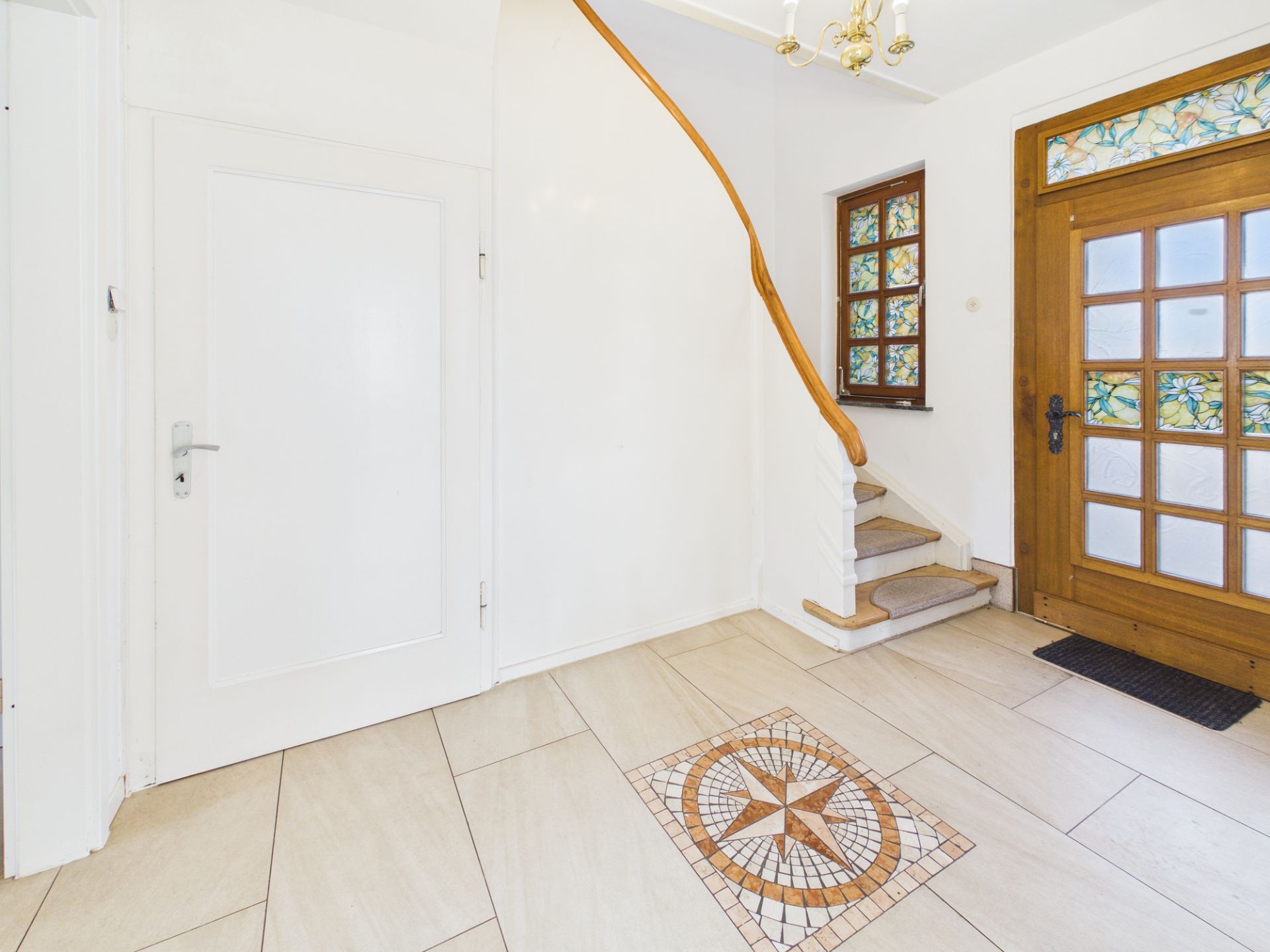 Heller Flur mit Holztür, Fenster, Fliesenboden und Treppe mit Holzgeländer.