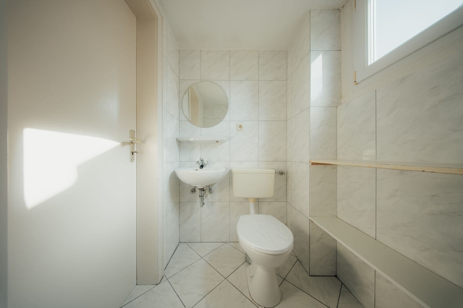 Helles Badezimmer mit Toilette, Waschbecken, rundem Spiegel und Fenster, weiße Fliesen an Wand und Boden.
